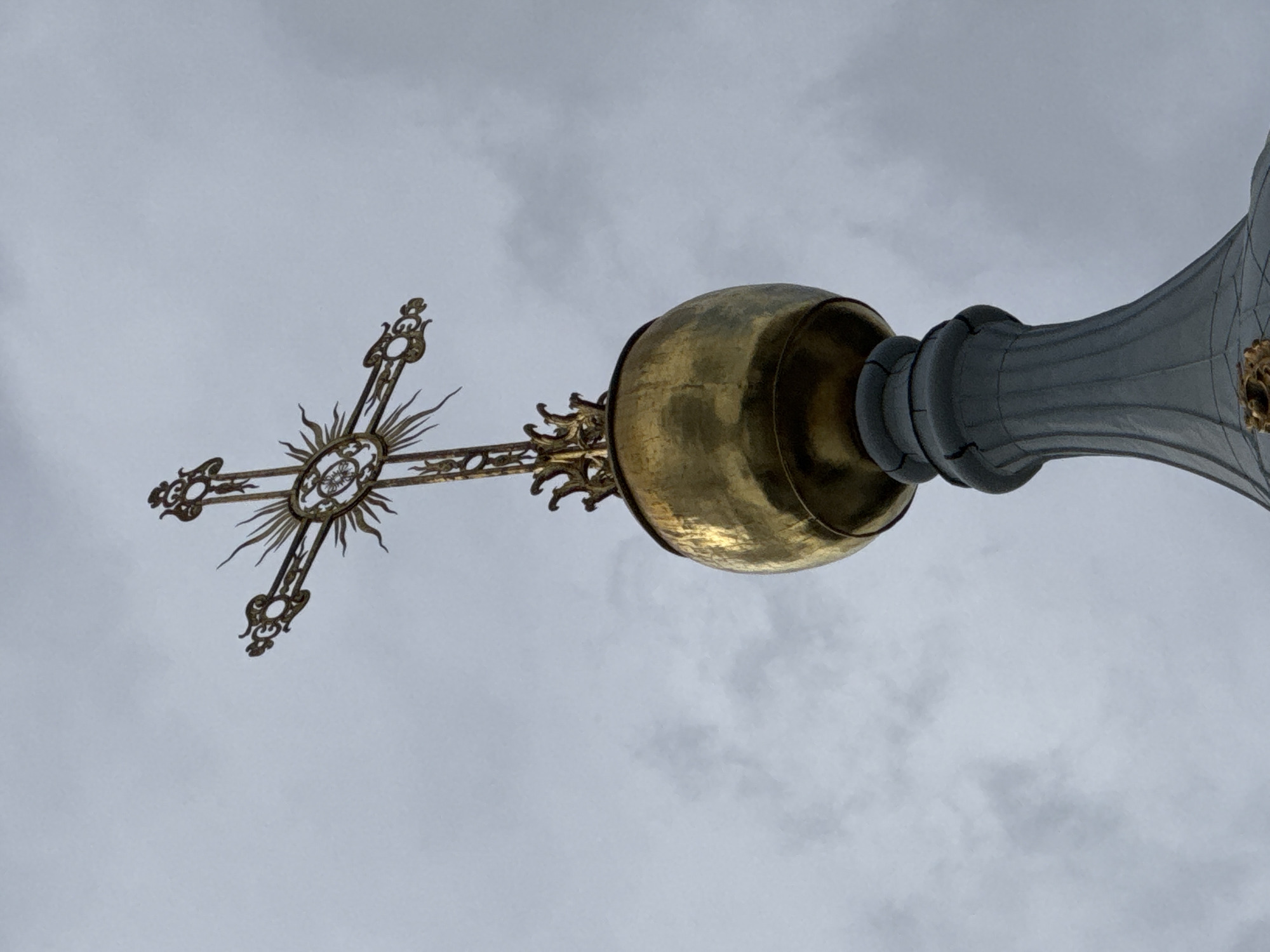 A gilded Orthodox cross adorns the top of a golden onion dome, which sits atop a grey, fluted architectural element. The structure is silhouetted against an overcast, cloudy sky. The image is a low-angle shot, looking up at the spire, with no people or further environmental details visible. The location is likely a church or cathedral in Saint Petersburg, Russia, given the architectural style.