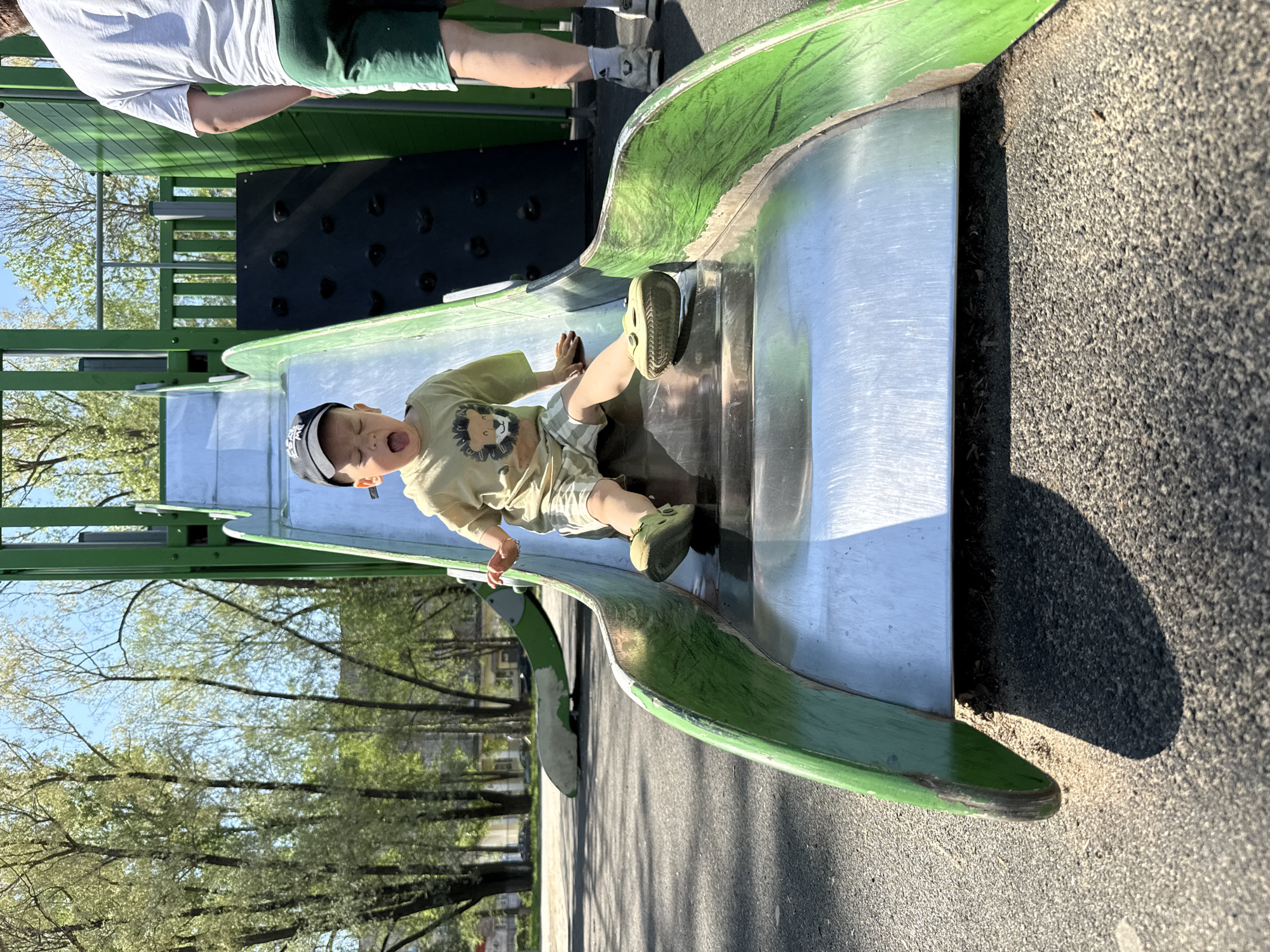 A child slides down a metal slide on a playground in Rusanovo, Russia. The child wears a baseball cap, a light-colored t-shirt with a lion graphic, patterned shorts, and green clogs. The child's mouth is open in an expression of excitement or surprise. The slide has green metal sides. An adult in a white t-shirt and green shorts stands at the top of the slide. In the background are trees, a climbing wall, and paved ground. The photograph is taken outdoors in daylight, with a shadow cast on the ground near the end of the slide.