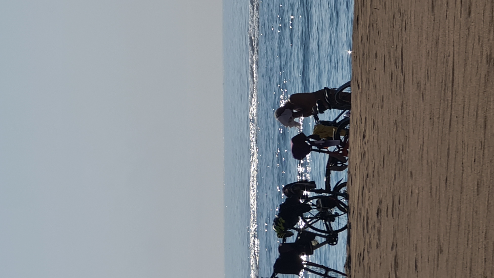In Denpasar, Indonesia, on a sandy beach, a person is seen drying themselves with a white cloth while standing next to a bicycle. The bicycle is equipped with panniers and a helmet, suggesting it is prepared for travel. In the background, the calm, sun-drenched ocean stretches to the horizon, with gentle waves lapping at the shore. The sky is clear and bright, indicating a sunny day.