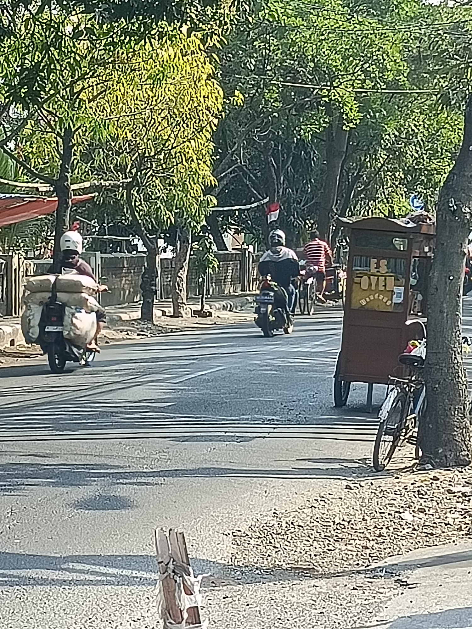 A sunny street scene in Kediri City, Indonesia, captures daily life with a focus on transportation and commerce. In the foreground, a motorcycle loaded with large sacks of goods is seen from the rear, with its rider wearing a white helmet. To the right, a mobile ice cart, painted brown and adorned with the text "ES OYEN," is parked near a tree. A bicycle is leaning against the tree. Further down the street, another motorcycle carries a rider with a red and white striped shirt, and behind him, another individual on a motorcycle is visible. Trees line the street, casting shadows on the asphalt road. The environment appears to be a typical urban setting with sidewalks and buildings in the background.