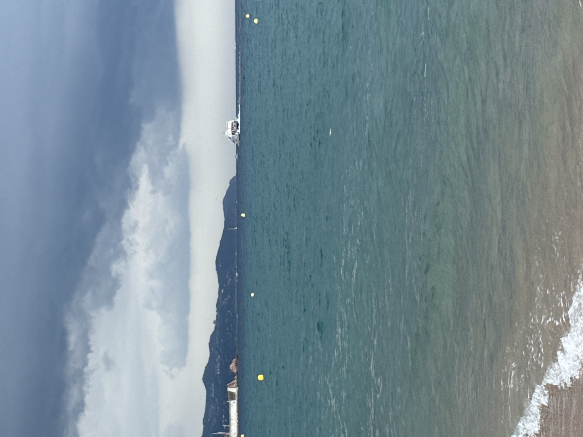 This is an eye-level, medium shot of a coastal scene in Blanes, Spain. The foreground features the edge of a sandy beach with gentle waves lapping at the shore, creating white foam. The water transitions from a light, sandy hue near the beach to a deep turquoise further out. Several yellow buoys are visible in the water. In the middle ground, a white ferry with red accents sails across the sea, leaving a wake behind it. In the background, a mountainous coastline stretches across the horizon, dotted with some buildings and trees. A large, dark grey cloud formation dominates the sky, suggesting overcast weather. A white structure with a red-tiled roof is visible on the left side of the coastline, possibly a lighthouse or a building. Sailboats are also visible in the distance on the sea.