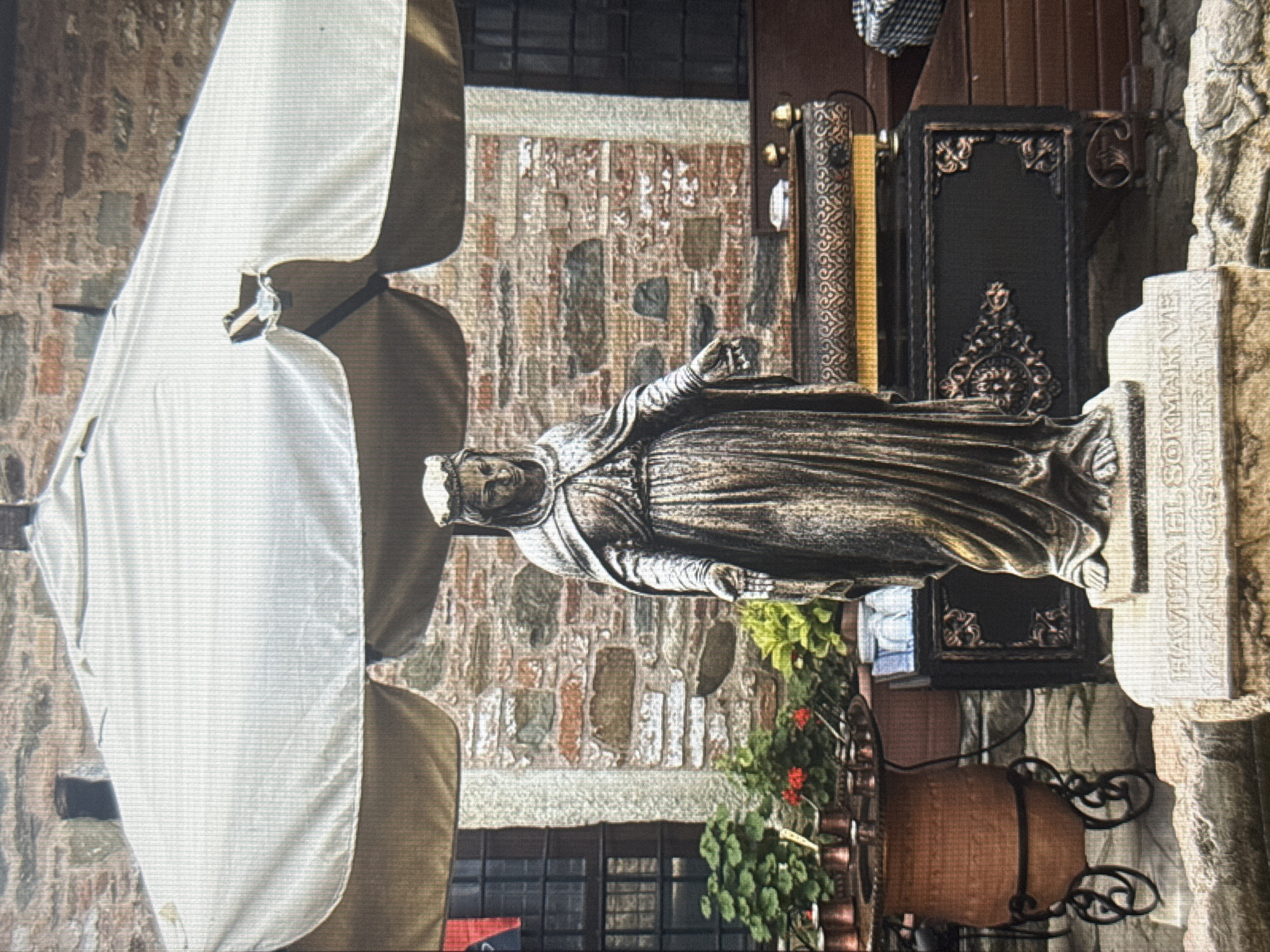 This image depicts an outdoor scene, possibly a cafe or courtyard, with a statue of a robed figure wearing a crown. The statue is made of a dark, metallic material and is positioned centrally. To the left, a large, white umbrella provides shade, its fabric slightly rumpled. Behind the statue, a rustic stone wall forms the backdrop, interspersed with dark wooden structures. A dark, ornate decorative book is visible to the right of the statue. Green plants with red flowers are growing in pots, adding a touch of color to the scene. A large terracotta pot with decorative metalwork sits in the foreground to the right. The overall lighting suggests daytime. Visible text is present on a stone slab to the far right, which reads "HAVUZA EL SORMAK VE FANCISCUS". The setting appears to be in Eyüpsultan, Turkey, suggesting a historical or cultural context.