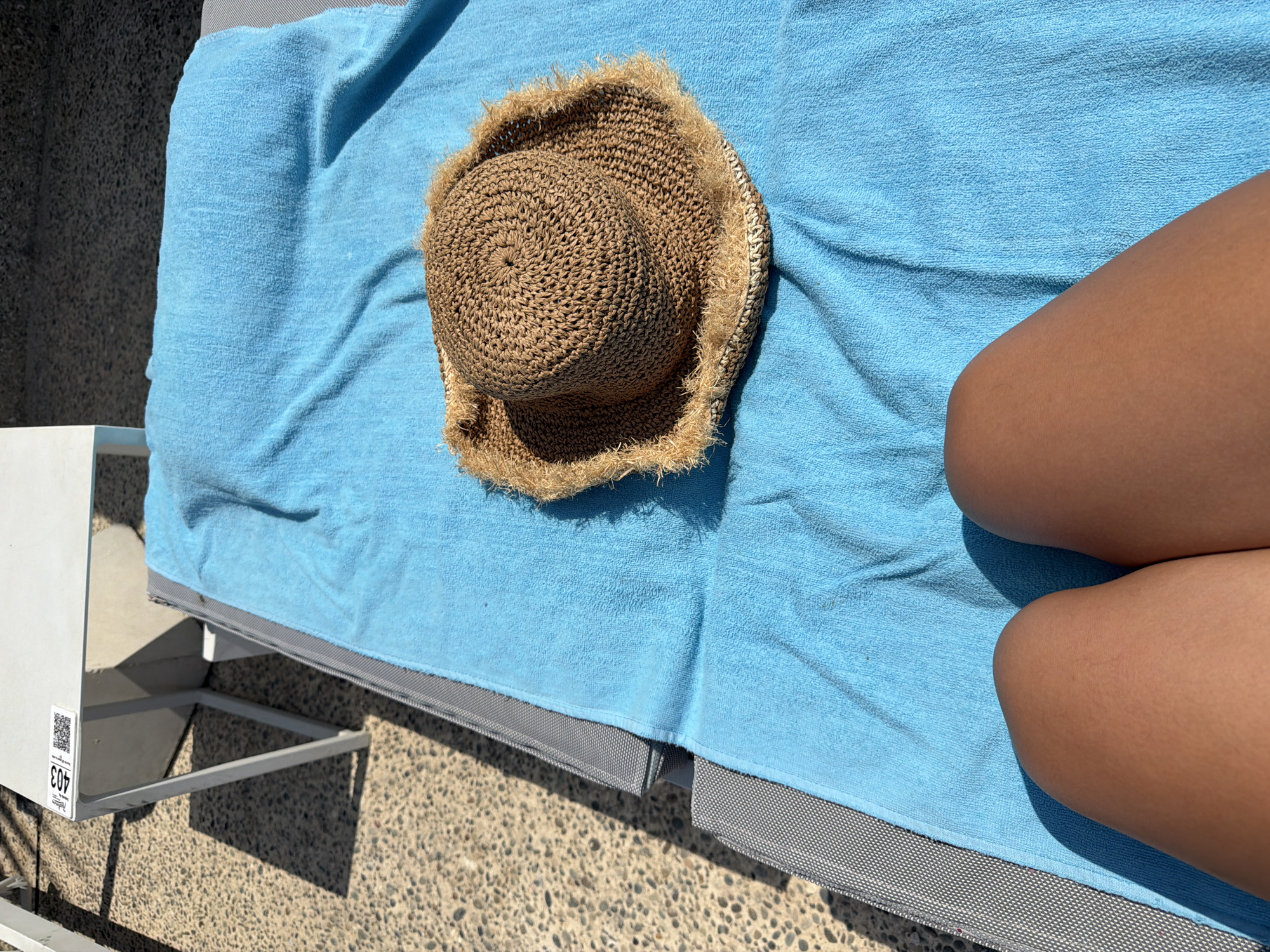An overhead, high-angle perspective shows a light brown, woven straw hat with frayed edges resting on a bright blue towel. The towel covers a grey lounge chair with a diamond-patterned fabric. The lounge chair is positioned on a speckled grey and tan paved surface. To the left of the lounge chair, a portion of a white table with a black and white barcode sticker is visible. The scene appears to be outdoors, likely in a public area or resort in Oroklini, Cyprus, given the context. The lower portion of the image shows a person's tanned legs with knees visible, positioned at the bottom of the frame, indicating they are sitting or lying on the lounge chair. The lighting suggests a sunny day with shadows cast on the paved surface.