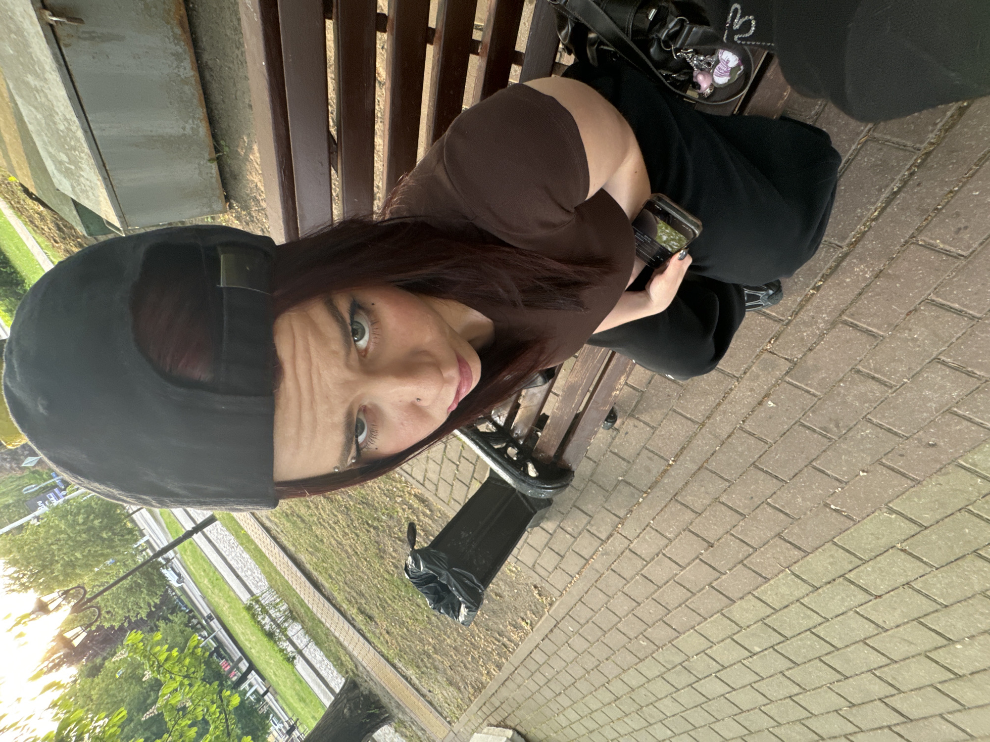 The image shows a woman sitting on a wooden bench, possibly in a park. She wears a brown top, black pants, and a black baseball cap. She is holding a smartphone in her hand and appears to be looking at the camera. Beside her on the bench is a black bag. A trash can is positioned near the bench. The area is paved with rectangular blocks and there's grass and trees in the background.