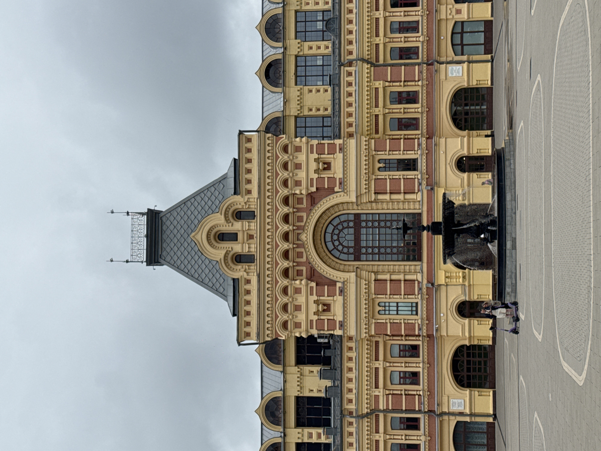The image depicts the Nizhny Novgorod Fair, a historic landmark in Nizhny Novgorod, Russia. The building features a grand, ornate facade with a combination of yellow and red brickwork, intricate decorative elements, and a distinctive mansard roof. A large, arched window dominates the central part of the building. In the foreground, a fountain is visible, and two individuals are present in the square, one of whom is standing next to a purple electric scooter. The sky is overcast, suggesting a cloudy day. The ground of the square is paved with cobblestones arranged in a patterned design.