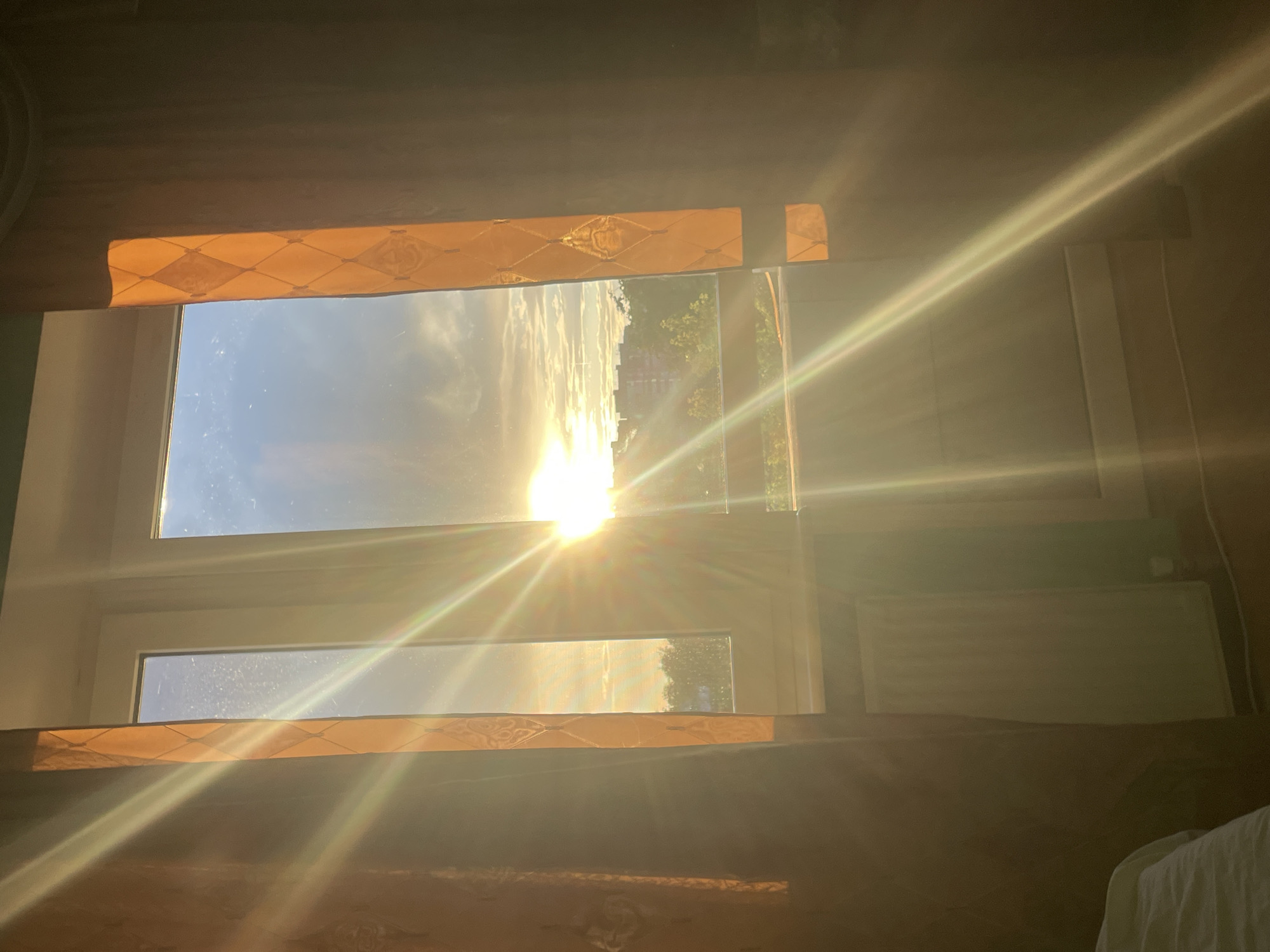 A view through a window frames a sunset with dramatic sun flares. The sun is low in the sky, positioned towards the right side of the window. Its bright rays create diagonal streaks of light that cut across the scene. Outside the window, a few buildings and trees are visible, silhouetted against the colorful sky. The window itself is a modern, white-framed structure, divided into two vertical panes. On either side of the window, dark curtains with an orange diamond pattern are partially drawn, with one curtain on the left appearing to be a dark brown or black. The foreground shows a radiator beneath the window. The overall impression is one of a warm, atmospheric evening, with the natural light creating a strong visual effect.