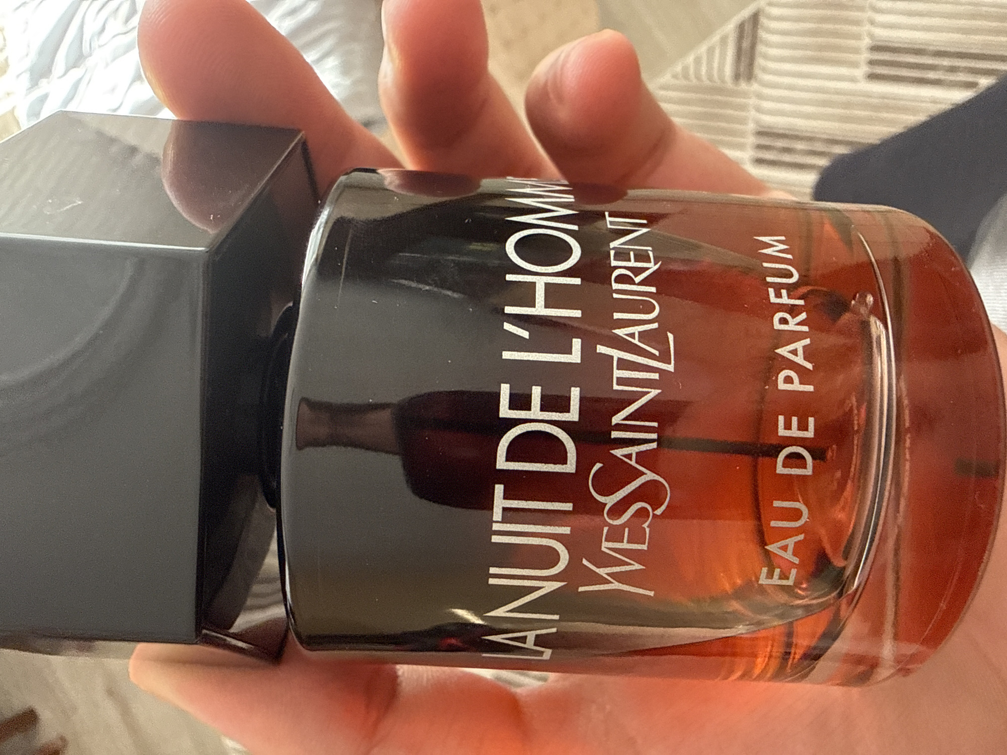 An extreme close-up shot shows a hand holding a bottle of Yves Saint Laurent "La Nuit de L'Homme" Eau de Parfum. The bottle is amber-colored and features white text. The background is out of focus and appears to be a striped surface. There is no indication of the location being Draginovo, Bulgaria.