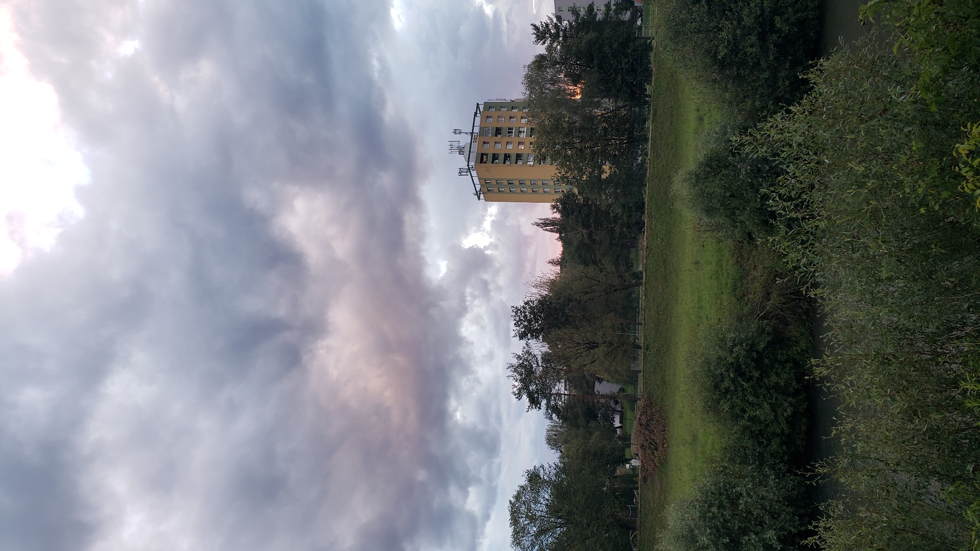 The image depicts a view of a riverbank in Prešov, Slovakia, with a tall, yellow apartment building visible in the background. The foreground features dense green bushes and vegetation along the river. Beyond the bushes, a grassy slope rises, dotted with trees. The apartment building, which appears to be around 10-12 stories high, is situated amidst a line of trees and some smaller buildings. The sky is filled with dramatic, grey clouds, with hints of pink and purple suggesting either sunrise or sunset. The overall scene conveys a somewhat overcast and tranquil atmosphere of a city outskirts.