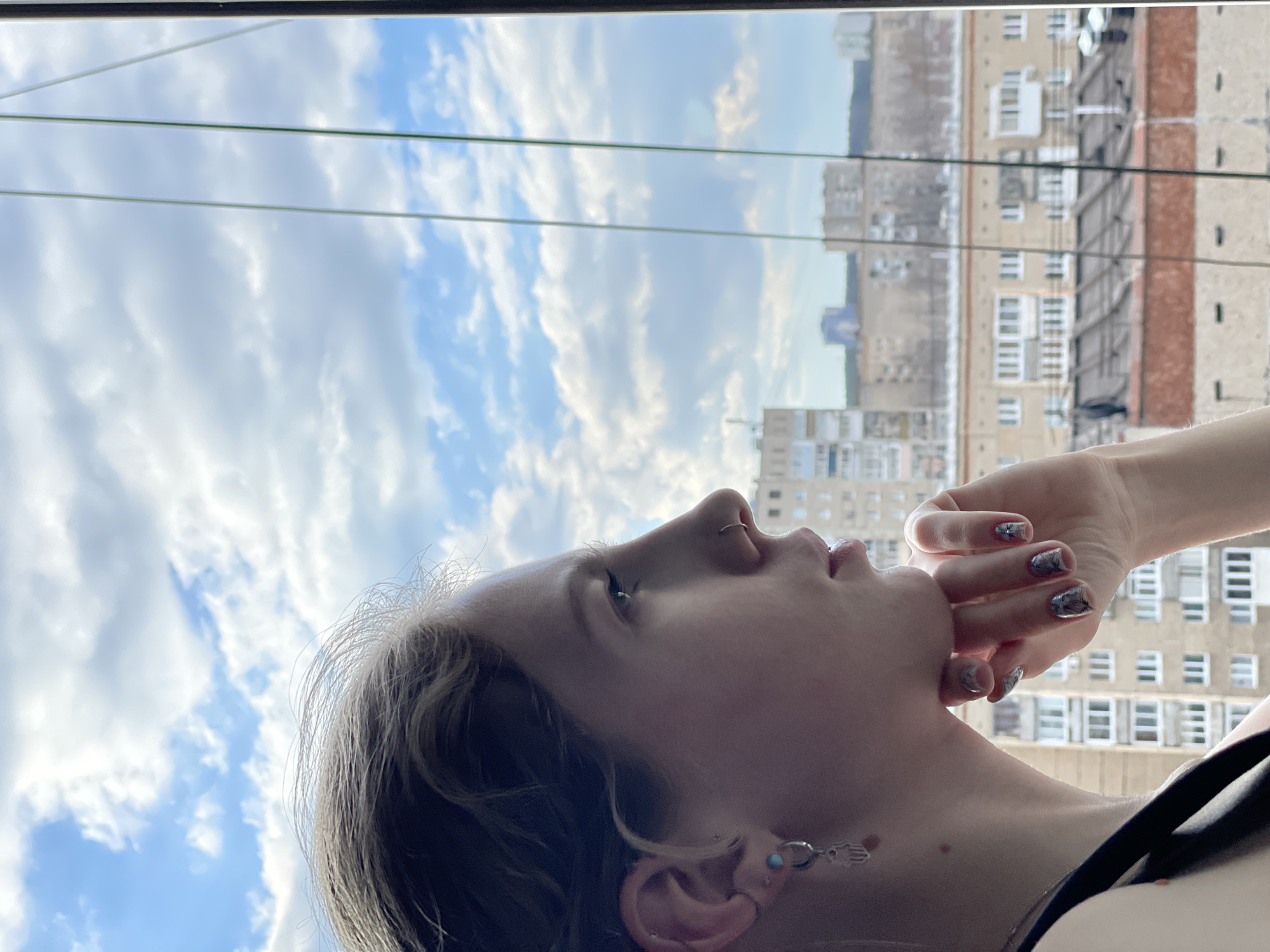 The image is a bright, upward-angled shot, rotated 90 degrees counter-clockwise, capturing a person looking skyward from an indoor setting in Izhevsk, Russia. The main subject, a person with light-colored hair, is seen in profile with their head tilted back, gazing towards the top of the frame. They wear a nose ring and an earring resembling a Hamsa hand on their left ear. Their right hand is positioned near their chin or mouth, showcasing manicured fingernails with a distinct, intricate pattern, possibly marbled or abstract.
The background features a expansive, cloudy sky with hints of blue, suggesting daytime. Several utility wires or cables run horizontally across the mid-ground, indicating an urban environment. Beyond these wires, multiple large, multi-story residential buildings with numerous windows are visible, hinting at the cityscape of Izhevsk. The lighting is bright and diffused, consistent with an overcast or partially cloudy day. No discernible text is present in the image. The overall scene conveys a moment of quiet observation or contemplation.