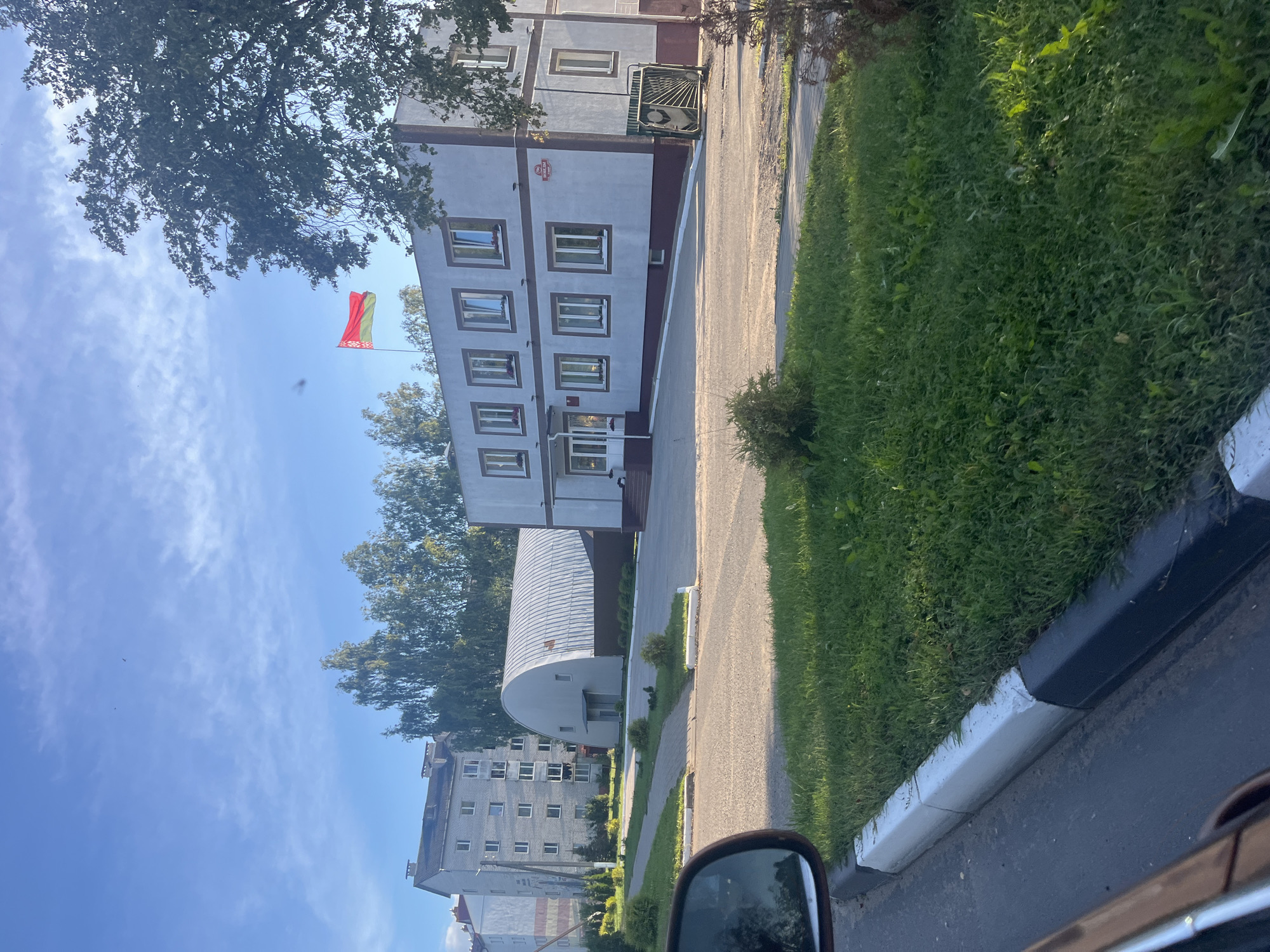 In the city of Lahojsk, Belarus, a building with a Belarus flag flying from its roof is visible on a sunny day. The building is light gray with dark gray trim and has multiple windows. To the left of this building, a larger, multi-story apartment building stands, also light gray. Between the two buildings, a large, arched hangar-like structure with a corrugated metal roof is present. The foreground shows a paved area with a grassy strip along the side, separated by a white and black painted curb. The scene appears to be a public or institutional area, possibly a school or administrative building complex, given the architectural styles and the presence of the flag. The sky is clear blue with scattered white clouds and a few birds are visible in the distance.