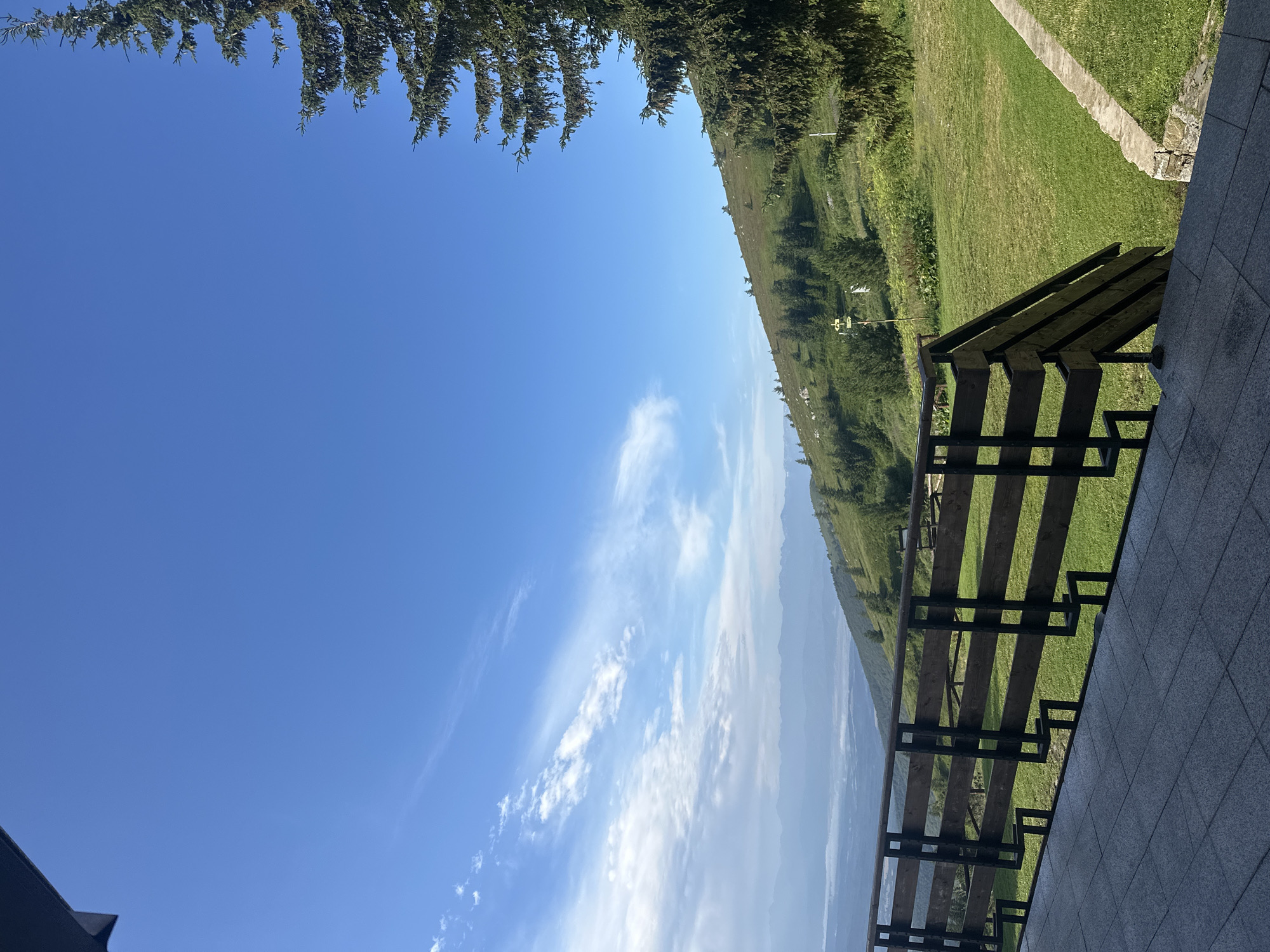 The image depicts a serene mountain landscape viewed from a tiled patio with a wooden railing. The sky is a vibrant blue, with wispy white clouds streaking across the upper left portion of the frame. A large evergreen tree dominates the right side of the image, its branches reaching towards the sky.
The foreground features a dark gray tiled patio leading to a wooden staircase with a dark metal railing. The railing is constructed of horizontal wooden planks supported by vertical metal bars, forming a geometric pattern. Beyond the patio, a lush green lawn slopes gently upwards towards a verdant hillside. The hillside is dotted with scattered trees and bushes, and a winding path or road is visible in the distance.
In the background, a range of mountains stretches across the horizon, their peaks softened by a light haze. The valley below appears misty, suggesting a significant elevation. A few small, weathered signs are visible on the hillside, one appearing to be a directional marker. The overall atmosphere is peaceful and natural, evoking a sense of tranquility and open space. The scene is likely a scenic overlook or a resting point within a mountainous area.
The location is identified as a spot in the city of Bistritsa, Bulgaria. This specific viewpoint is likely from a mountain lodge, a guesthouse, or a scenic viewpoint located within the Vitosha Mountain range, which encompasses Bistritsa. The architectural elements like the patio and railing suggest a man-made structure designed to offer comfortable viewing of the natural surroundings.