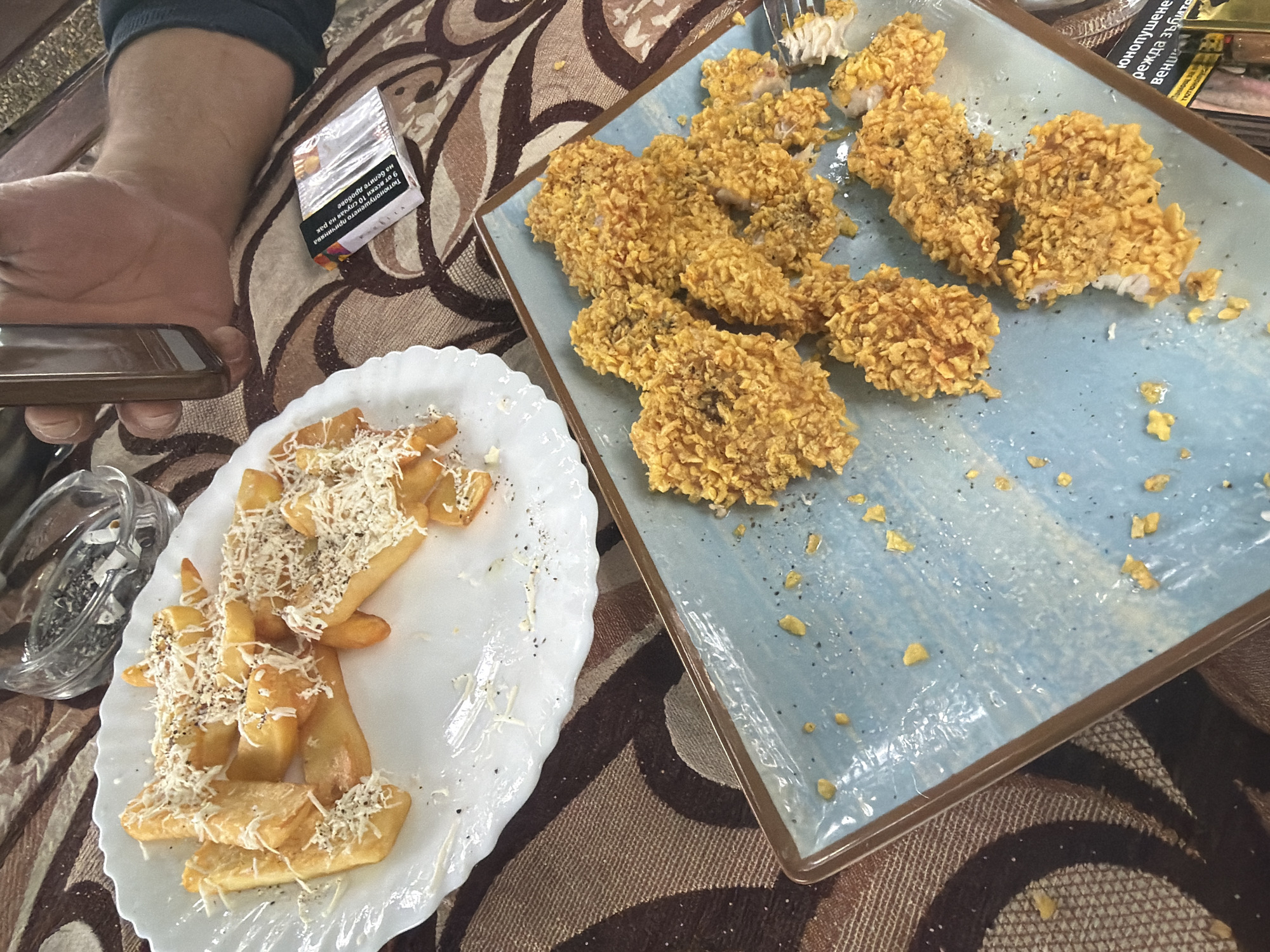 An overhead shot captures a casual dining scene in Sushina, Bulgaria. The image features a table set with two plates of food, a person's hand holding a smartphone, a cigarette pack, and an ashtray, all resting on a patterned tablecloth in brown and beige tones.
On the right, a rectangular light blue plate holds numerous pieces of fried chicken, likely nuggets or tenders, coated in a crunchy, golden, cereal-like breading. Some pieces are sprinkled with black pepper. One piece appears to have been partially eaten, revealing white meat inside. Scattered crumbs of the breading are visible on the plate.
To the left, a white oval plate with a scalloped edge contains thick-cut french fries or potato wedges, generously topped with shredded white cheese and black pepper. Nearby, a glass ashtray holds cigarette butts and ash.
A person's arm and hand are visible on the far left, holding a dark-colored smartphone, suggesting they are either about to eat, taking a photo, or engaging with their device during the meal. A partially obscured pack of cigarettes with Cyrillic text is also on the table.
Visible text on the cigarette pack includes health warnings such as "юнопушенето причинява рак на белите дробове" (smoking causes lung cancer) and mentions "режда зъбите" (damages teeth) and "вени" (gums), reinforcing the Bulgarian context. The bright lighting suggests it is daytime. The overall scene depicts a relaxed meal, possibly at a cafe or restaurant.