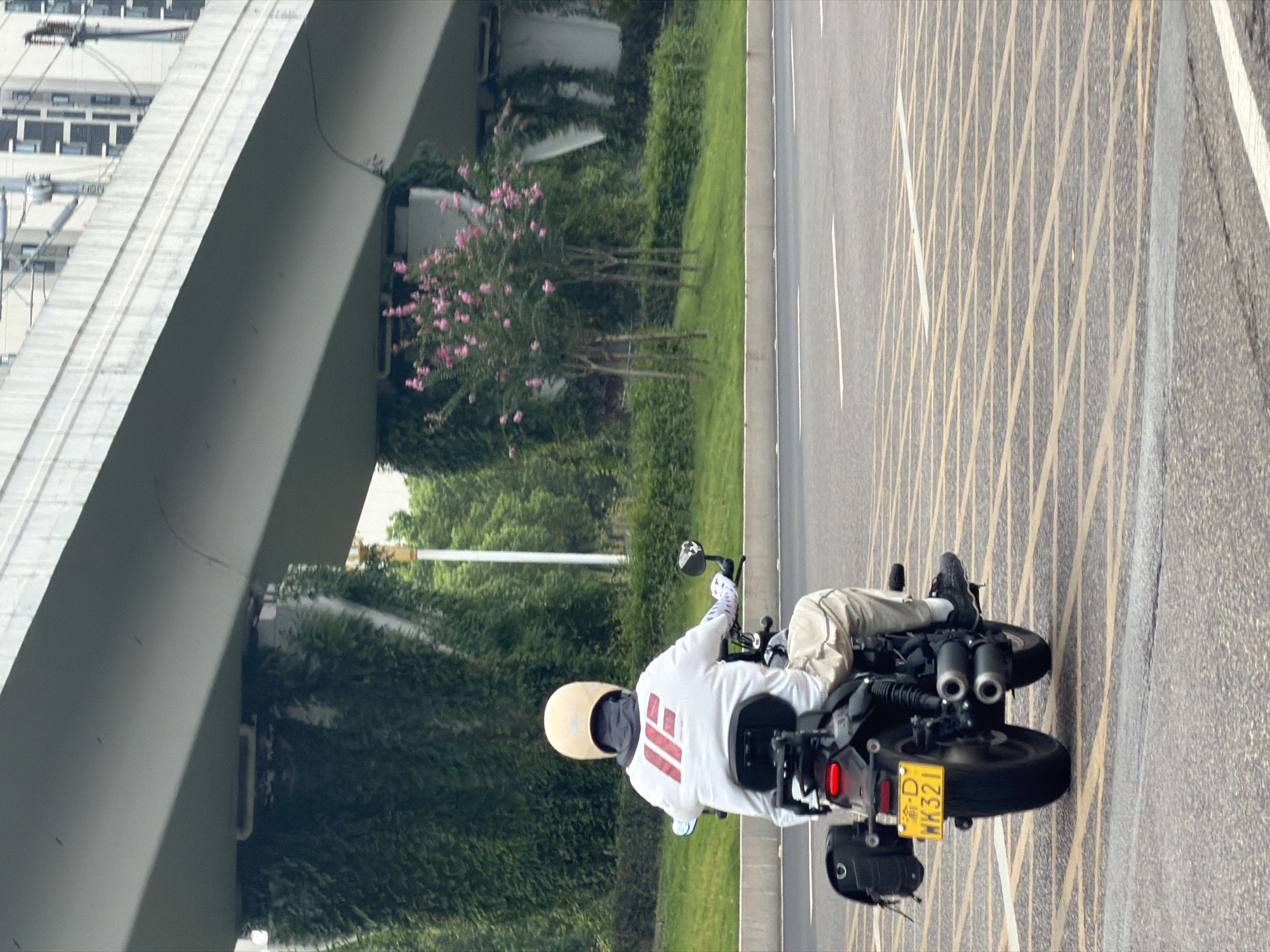 A rear view of a person riding a motorcycle on an asphalt road in Jiangjin, China. The rider is wearing a white long-sleeved shirt with red and white graphic elements, tan pants, and a light-colored helmet. The motorcycle is black with a black seat, a black bag attached to the left side, and a yellow license plate with Chinese characters and the alphanumeric sequence "WK321". The road has yellow grid markings and white lane lines. In the background, there is a large elevated concrete structure, likely an overpass, with thick vegetation growing on its support pillars. Trees with small pink flowers are also visible in the background. The sky is overcast.