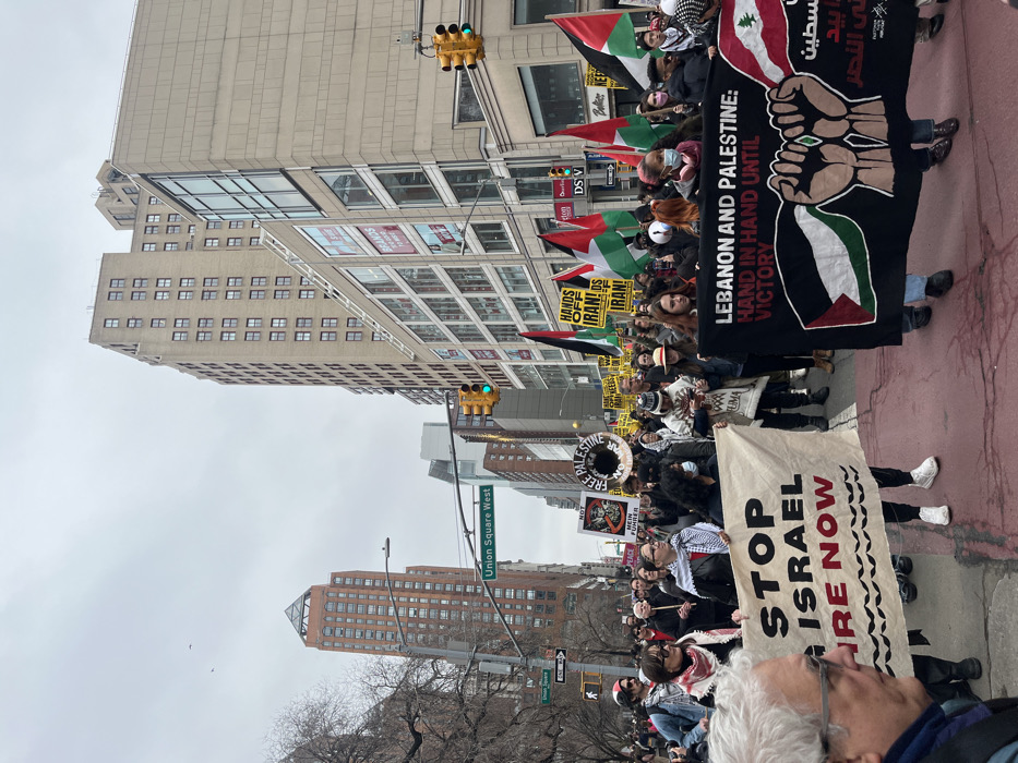 A street protest is depicted on Union Square West in New York City, United States of America, under an overcast sky. The roadway is occupied by a large gathering of individuals. In the foreground, a white banner is held horizontally, displaying 'STOP ISRAEL FIRE NOW' in black and red text, with a wavy black design below. Adjacent to this, a large black banner is carried, reading 'LEBANON AND PALESTINE: HAND IN HAND UNTIL VICTORY', which features two clasped fists, a Palestinian flag on the left, a Lebanese flag on the right, and Arabic script. Numerous participants in the mid-ground display Palestinian flags (black, white, green, red triangle) and black flags. Several yellow signs are visible among the crowd, many stating 'HANDS OFF IRAN!'. Other discernible signs include a circular white sign reading 'FREE PALESTINE' and a white sign with 'PEACE'. The background features multi-story urban architecture, including a reddish-brown building with a pyramidal roof on the left and a tall, beige-colored building with numerous windows on the right. A green street sign explicitly marks the location as 'Union Square West'. Traffic signals are visible overhead, with one indicating a green light. The immediate road surface in the foreground is reddish-brown.