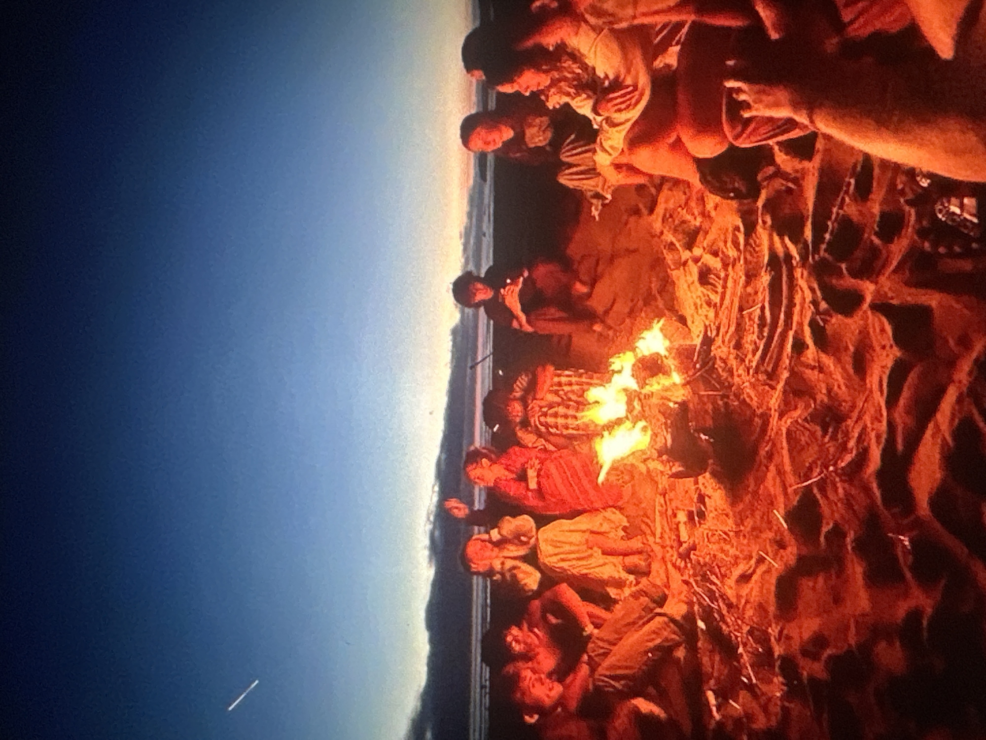 The image depicts a group of young people gathered around a bonfire on a beach at dusk. The sky is a gradient of deep blue to pale orange, with a faint contrail visible in the upper left quadrant. The fire casts a warm, orange glow on the sand and illuminates the faces of the individuals. Several people are seated in a circle, some facing the fire, others interacting with each other. Their clothing appears casual, consistent with a beach setting. The sand is textured and appears damp in places, suggesting proximity to the water. The background shows a dark silhouette of the ocean and possibly a coastline. The scene conveys a sense of casual gathering and social interaction in an outdoor, natural environment. There is no identifiable information within the image that suggests a specific location in Tallinn, Estonia.