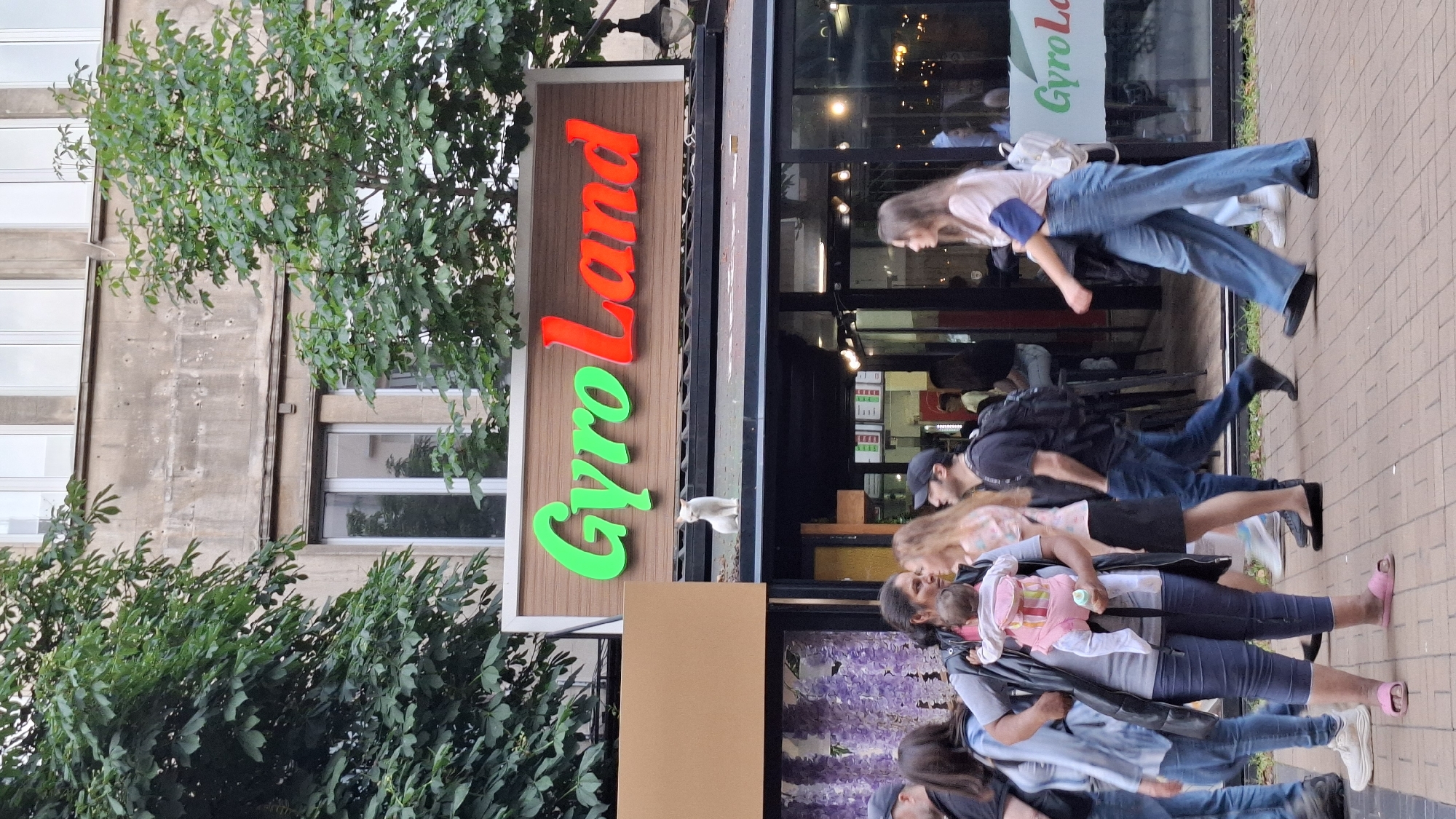 This image captures a street scene in Sofia, Bulgaria, featuring the "Gyro Land" restaurant, likely located at ul. "Georgi S. Rakovski" 105. The upper part of a light-colored building, exhibiting multiple small indentations or marks on its facade, is visible, partially obscured by green leafy trees. Below, the restaurant's sign displays "Gyro" in green illuminated letters and "Land" in red illuminated letters on a brown wooden background. Directly beneath the sign, on a black awning, a white cat is seated centrally, looking forward.
Below the awning, the restaurant's glass storefront reflects the surroundings and reveals interior lighting. In the foreground, on a brick-paved sidewalk, several individuals are walking from left to right. A woman in the center foreground wears a grey t-shirt, a dark vest, blue capri pants, and pink slide sandals. She carries an infant in a pink and multi-colored striped front-facing baby carrier; the infant, dressed in white and pink, holds a green bottle. To her right, a woman with light hair, wearing a patterned top and dark skirt, walks ahead. Behind them, a man with a dark t-shirt, black baseball cap, and black backpack also walks to the right. Further to the right, a woman wearing a light-colored long-sleeved top, wide-leg blue jeans, and a white backpack walks in the same direction. A partial view of a black ornate street lamp is visible on the far right edge of the frame.