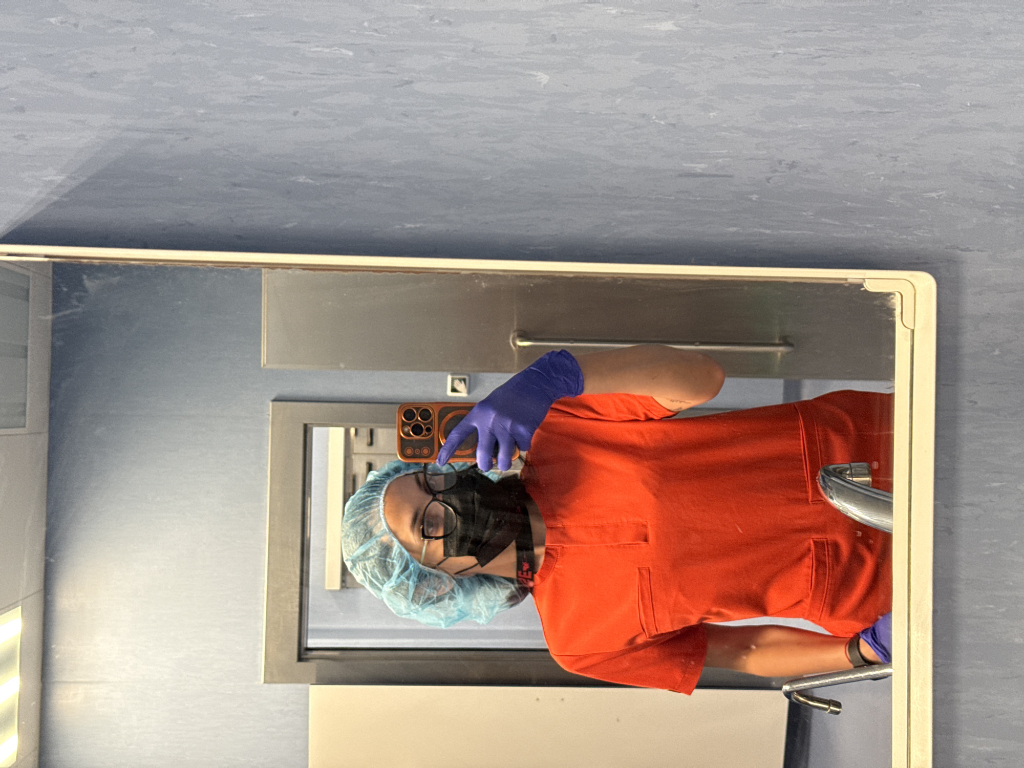 A person wearing medical attire, including a hair net, a face mask, and gloves, is taking a selfie in a bathroom mirror. The person is wearing an orange scrub top and is holding a cell phone in their left gloved hand. The bathroom has blue walls and stainless-steel fixtures, including a sink and cabinets. The reflection indicates a modern, possibly clinical, setting. There is no information available to indicate the location in Pleven, Bulgaria.