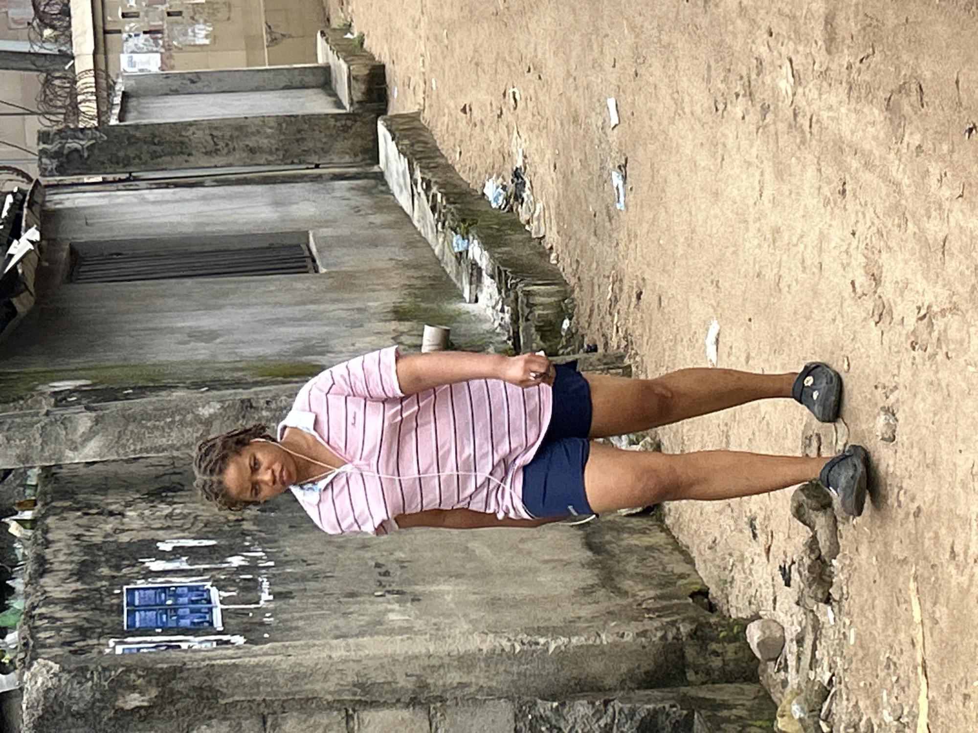 A woman with braided hair stands in an outdoor, urban setting in Ihiagwa, Nigeria. She is wearing a pink and white striped short-sleeved shirt and dark blue shorts. White earbuds are in her ears, with the cord visible around her neck. Her right hand is raised towards her face, and she is looking slightly downwards and to her left, with a neutral expression. She is wearing dark shoes.
The setting appears to be a rough, outdoor area with concrete structures and a dirt or sandy ground. There is visible debris and some vegetation growing around the edges of the concrete. A building with a barred window is visible in the background to the left. The image has a grainy texture, and the lighting suggests it was taken during the daytime, possibly under overcast conditions due to the diffused light. The overall impression is of a casual, everyday moment in a potentially underprivileged or developing area. There is no visible text in the image.