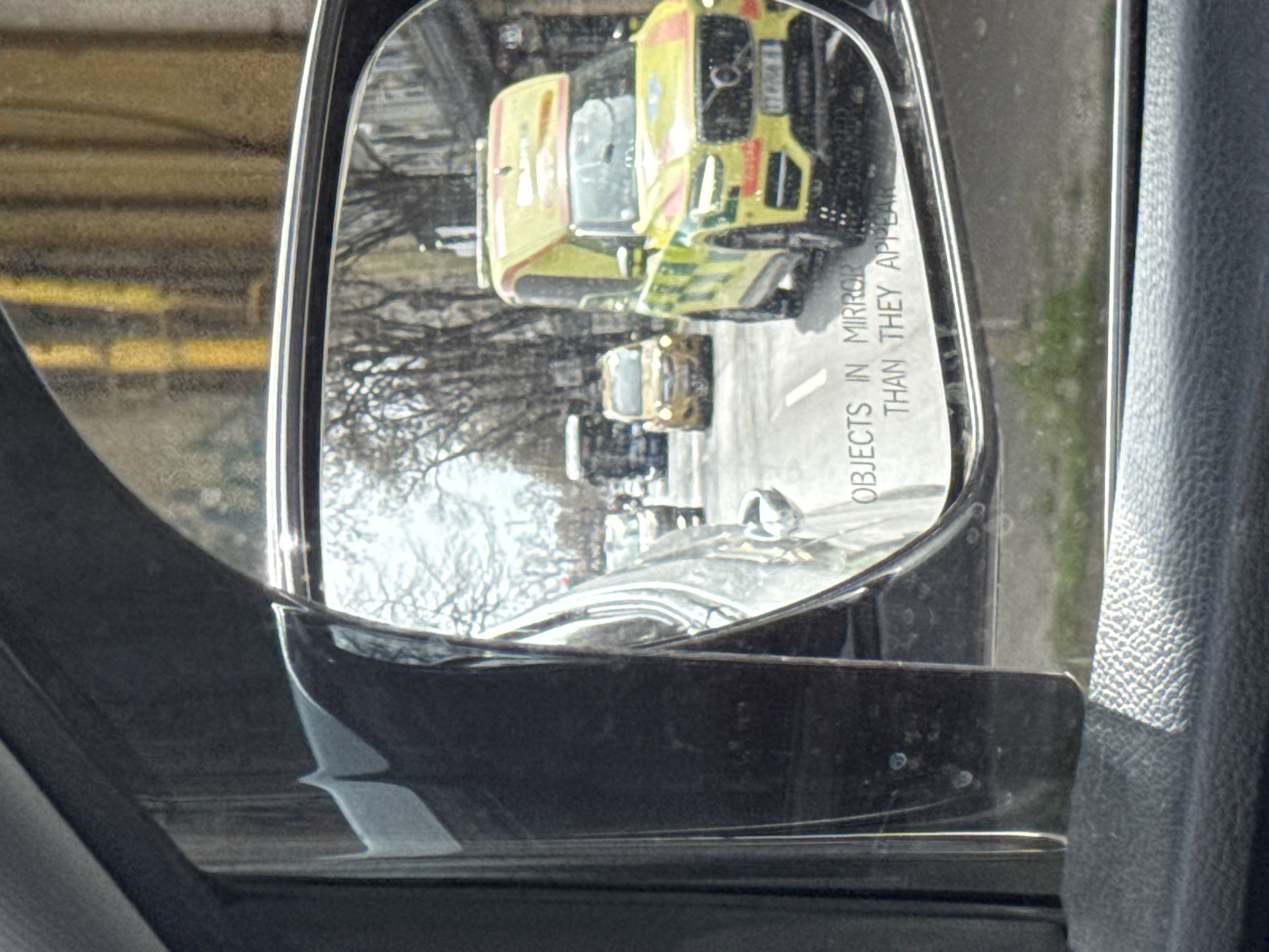 The image shows a side-view mirror of a car reflecting an ambulance in Varna, Bulgaria. The reflection also includes other cars, bare trees, and a street with white lines. The mirror includes the warning "OBJECTS IN MIRROR ARE CLOSER THAN THEY APPEAR." The interior of the car, including parts of the door and upholstery, is visible around the mirror. The environment appears to be a street in an urban area.