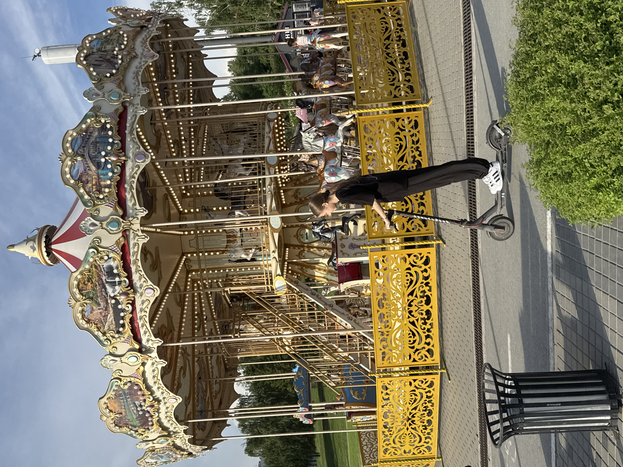 A young woman in black pants and a black t-shirt rides a black electric scooter past a yellow ornate fence. She is wearing white Adidas sneakers with black stripes. Behind the fence is a carousel with an ornate top decorated with paintings and lights, and carousel horses. The carousel is surrounded by trees. In the foreground, to the left of the woman, is a black trash can. The ground is paved with gray square tiles and asphalt. The sky is blue with some clouds. The scene is outdoors on a sunny day. The location is the Carousel "The Fairy Tale" in the Central Park of Culture and Recreation named after Kirov, in Novosibirsk, Russia.