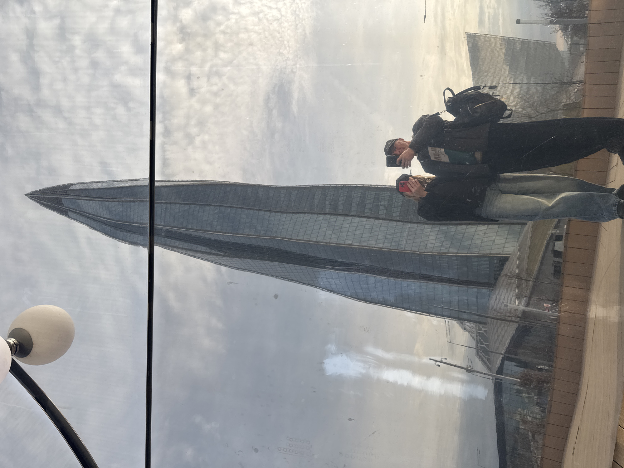 A reflection shows a couple taking pictures in front of the Lakhta Center in Saint Petersburg, Russia. The building's spire dominates the reflection, which also captures an overcast sky and part of the surrounding urban environment. The couple is standing on a paved surface, both holding smartphones pointed towards the skyscraper. One person is wearing a backpack. A streetlight fixture is visible on the left edge.