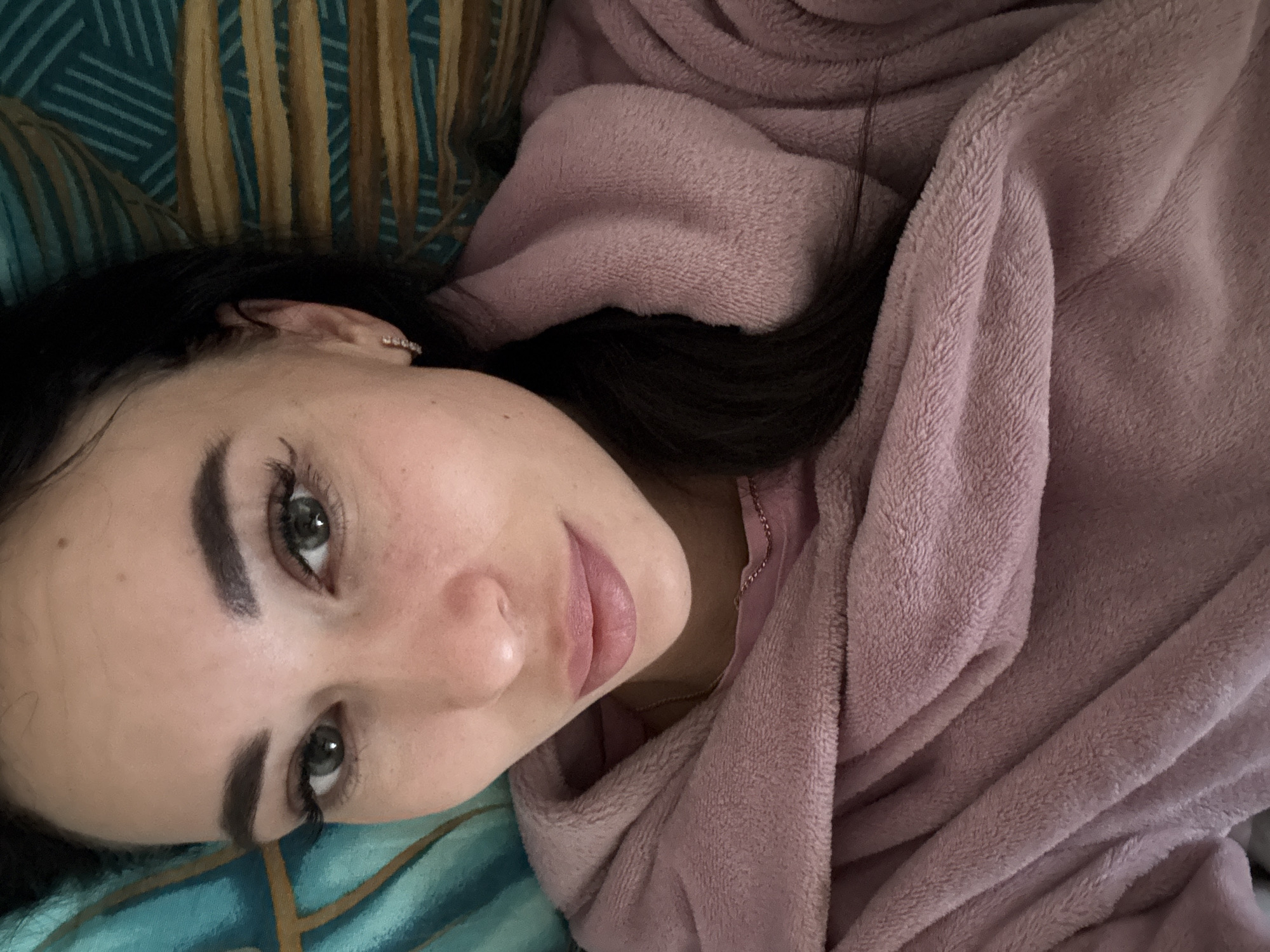 A young woman with dark hair and fair skin is lying down. Her head rests on a patterned blue and yellow surface. She is partially covered by a light pink, soft, textured robe. Her eyes are gray-green. She has dark eyebrows and full lips. A piece of jewelry is visible in her left ear. A fine gold chain is around her neck. The scene appears to be indoors. There is no information to infer that the image was taken in the city of Chaikovsky, Russia.