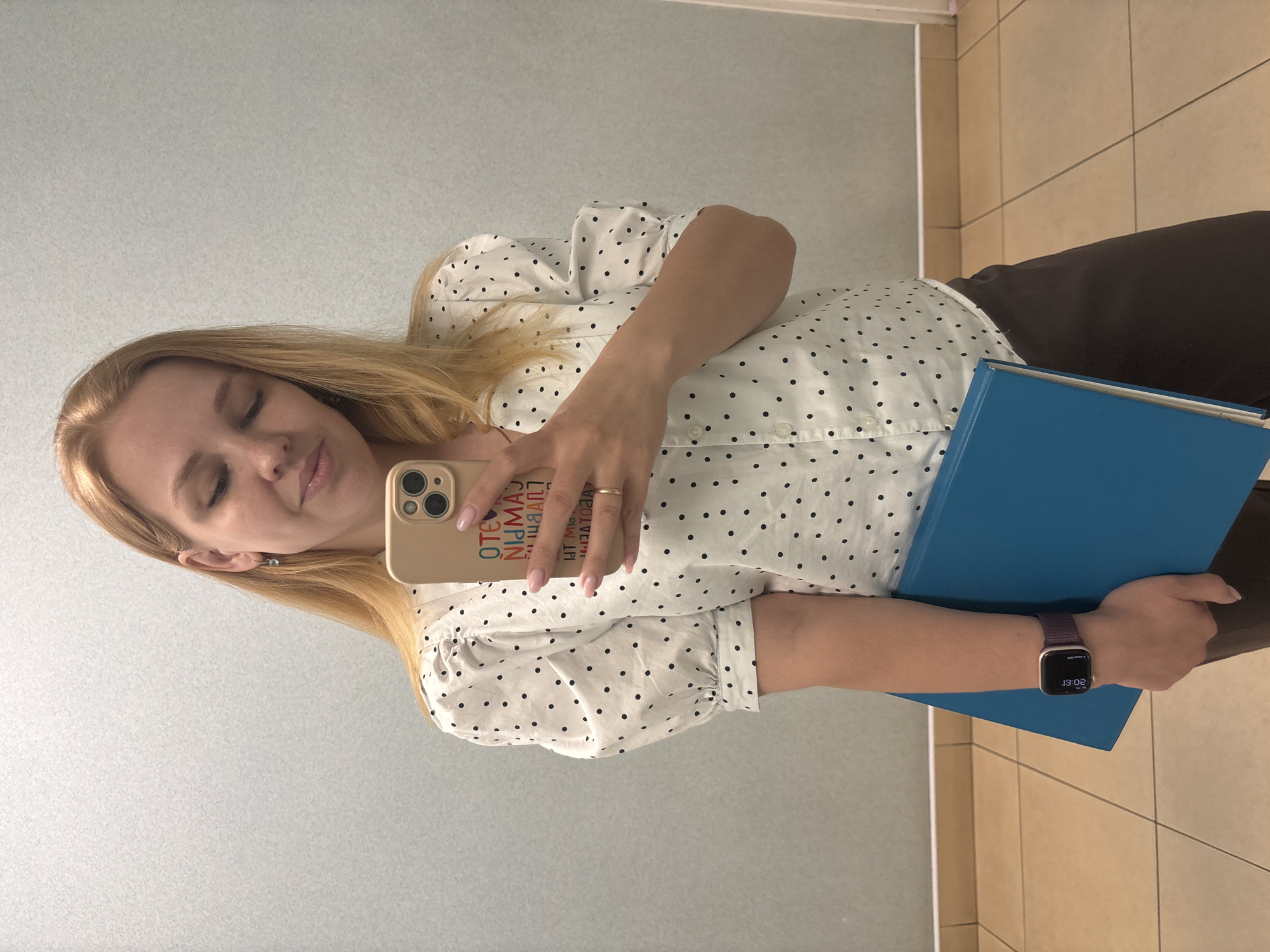 A young woman with blonde hair stands facing a mirror, taking a selfie. She is wearing a white blouse with black polka dots, dark trousers, and a wristwatch. She is holding a blue folder in her left arm. The background appears to be an indoor setting, possibly a restroom or a locker room, with tiled floors and paneled walls. There are no discernible features that would place the image specifically in Шебунино, Russia.