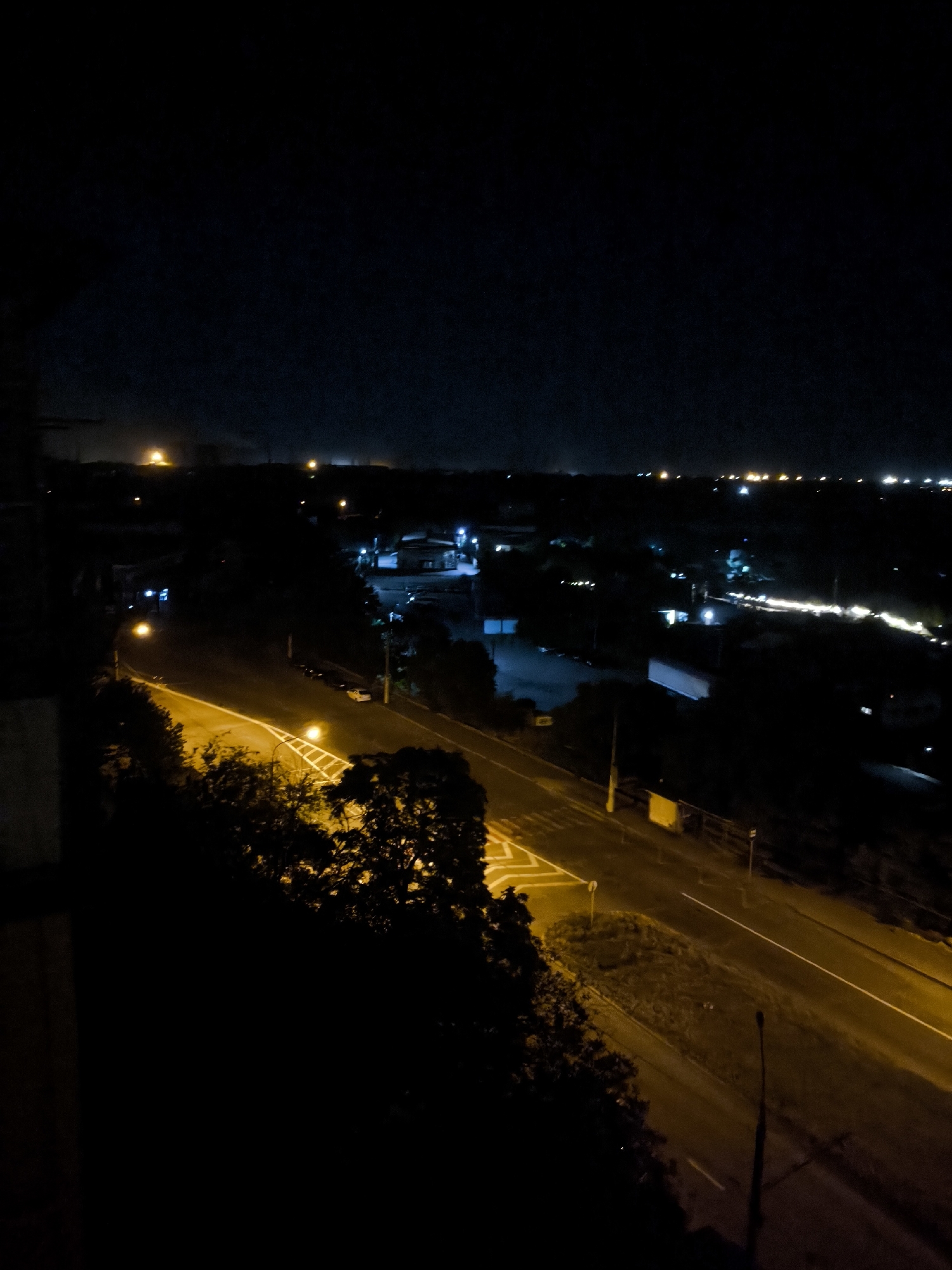 The image captures a nighttime view of a street and surrounding cityscape, likely in Zaporizhzhia, Ukraine, given the provided context. The foreground is dominated by dark foliage and a building structure on the left, obscuring a significant portion of the scene. A well-lit street, illuminated by streetlights and the headlights of a vehicle, traverses the mid-ground diagonally from left to right. The street appears to have multiple lanes and is marked with white lines. The background reveals a spread of city lights and structures receding into the darkness, suggesting an urban environment. The overall impression is one of a quiet, dimly lit evening in a city, with the artificial lights of the street and buildings cutting through the natural darkness.