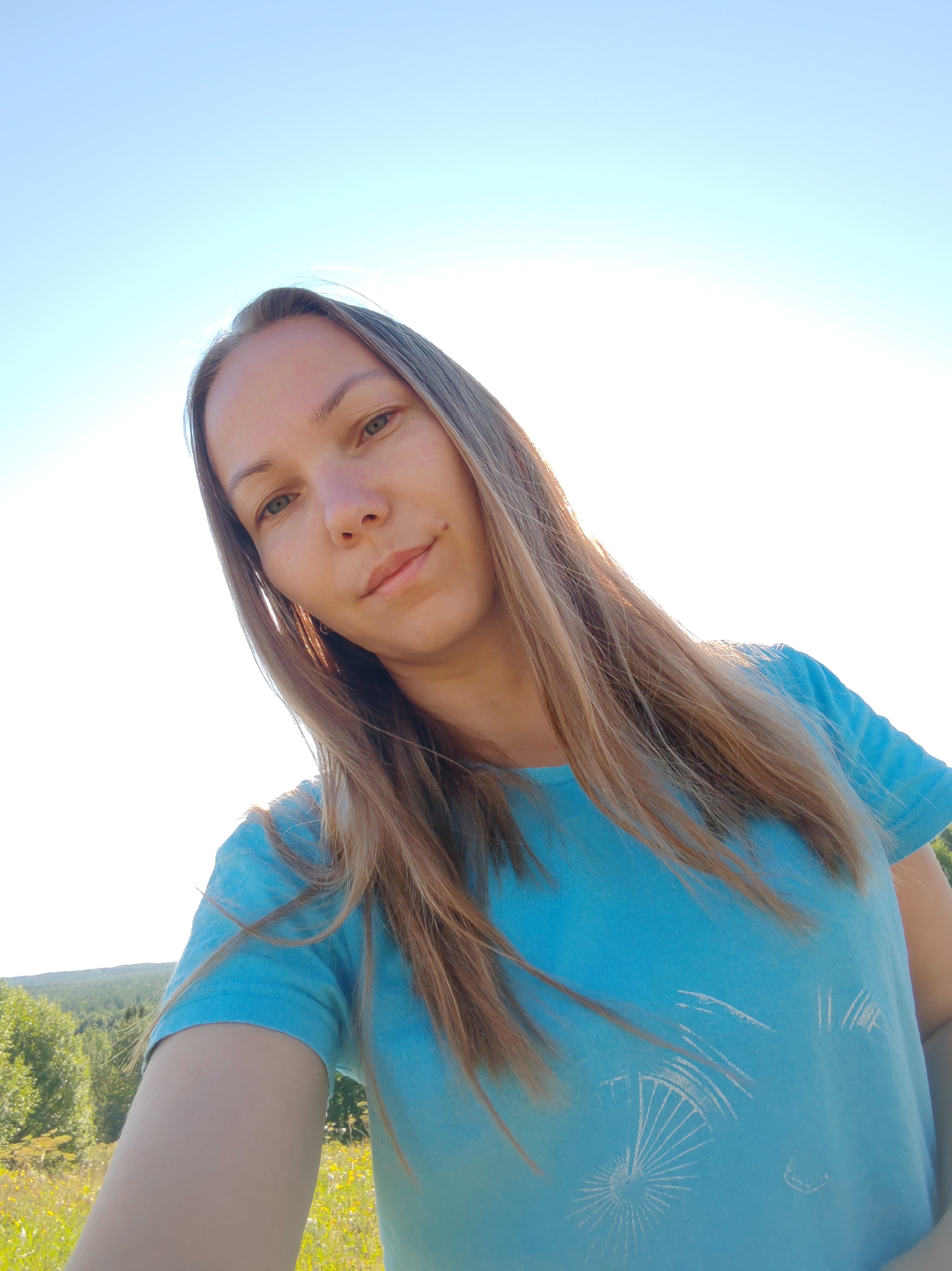 A young woman with long, light brown hair and green eyes is taking a selfie outdoors. She is wearing a bright blue t-shirt with a white graphic of a bicycle on it. The background shows a clear blue sky and a green, hilly landscape with trees and wildflowers. The sun is bright and appears to be setting or rising, casting a warm glow on her face and hair. The scene suggests a peaceful and natural setting, possibly in a rural area like Чухлэм, Russia, given the natural landscape.
