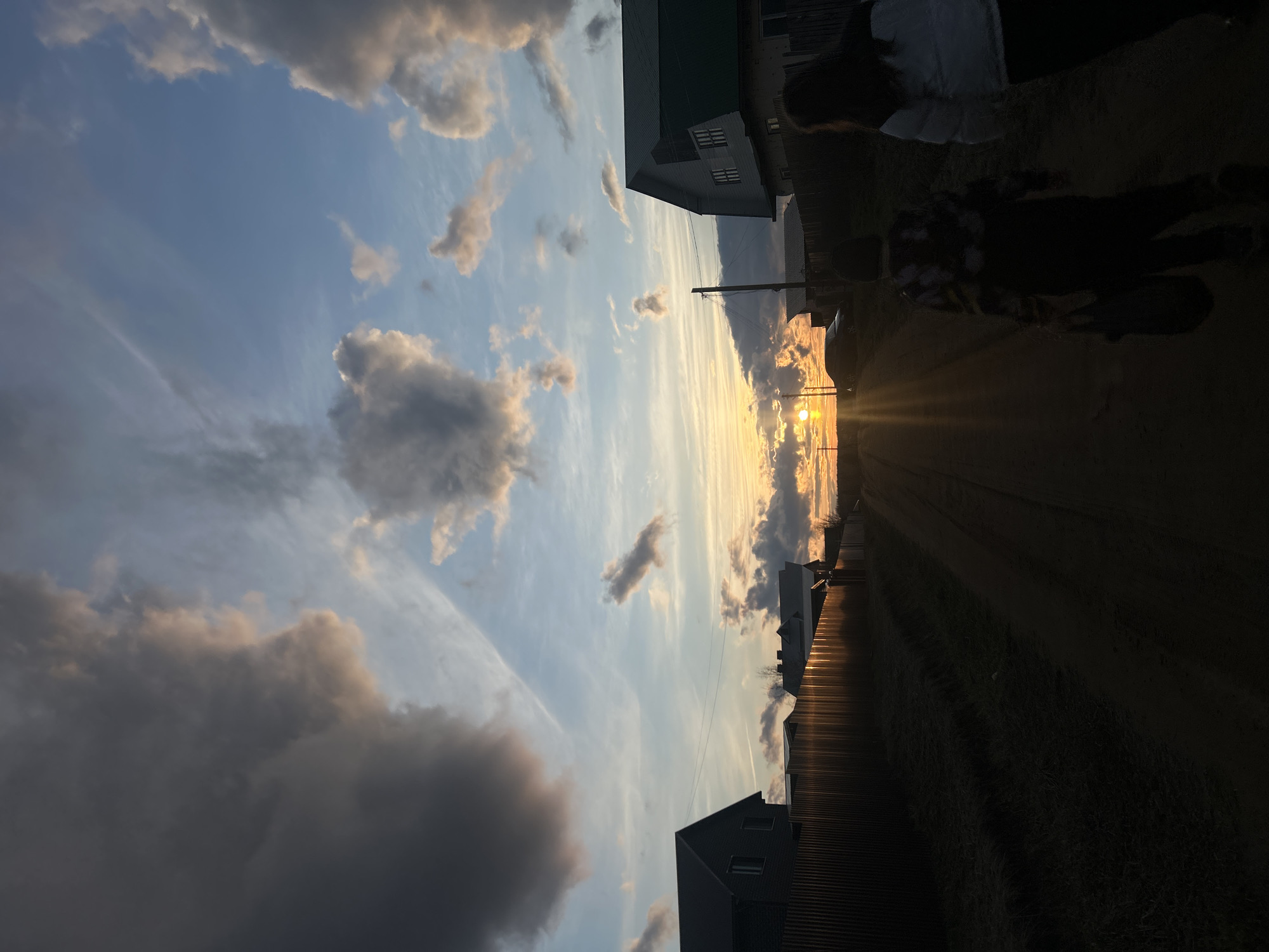 The image captures a sunset scene in what appears to be a rural or semi-rural settlement, consistent with the provided location of Olkhovka, Russia. The sky occupies a significant portion of the frame, displaying a gradient from light blue overhead to warm yellow and orange hues near the horizon, where the sun is brightly visible, casting a strong glare. Various clouds are scattered across the sky; some are dark grey and heavy, while others are illuminated by the low sun, showing warm orange and pink tones. Wispy, cirrus-like clouds also streak across the blue expanse.
A dirt or unpaved road runs through the center-right of the image, leading towards the vanishing point where the sun is setting. The sides of the road are lined with fences and buildings. On the left side, a house with a dark roof and visible windows is partly seen in the foreground, adjacent to a corrugated metal fence. Further along, other houses, including one with a green roof, are visible. On the right side, the foreground is heavily shadowed, almost entirely in silhouette, due to the direct light from the setting sun.
Two human figures are discernible in silhouette on the right side of the road; one appears to be an adult, and the other is shorter, possibly a child or another adult. They are standing or walking towards the horizon. A utility pole stands mid-ground on the right, and a car is partially visible in silhouette behind the figures. The overall atmosphere suggests a quiet evening, with the warm, dramatic light of the sunset illuminating the sky and casting long shadows across the landscape. No visible text is present.