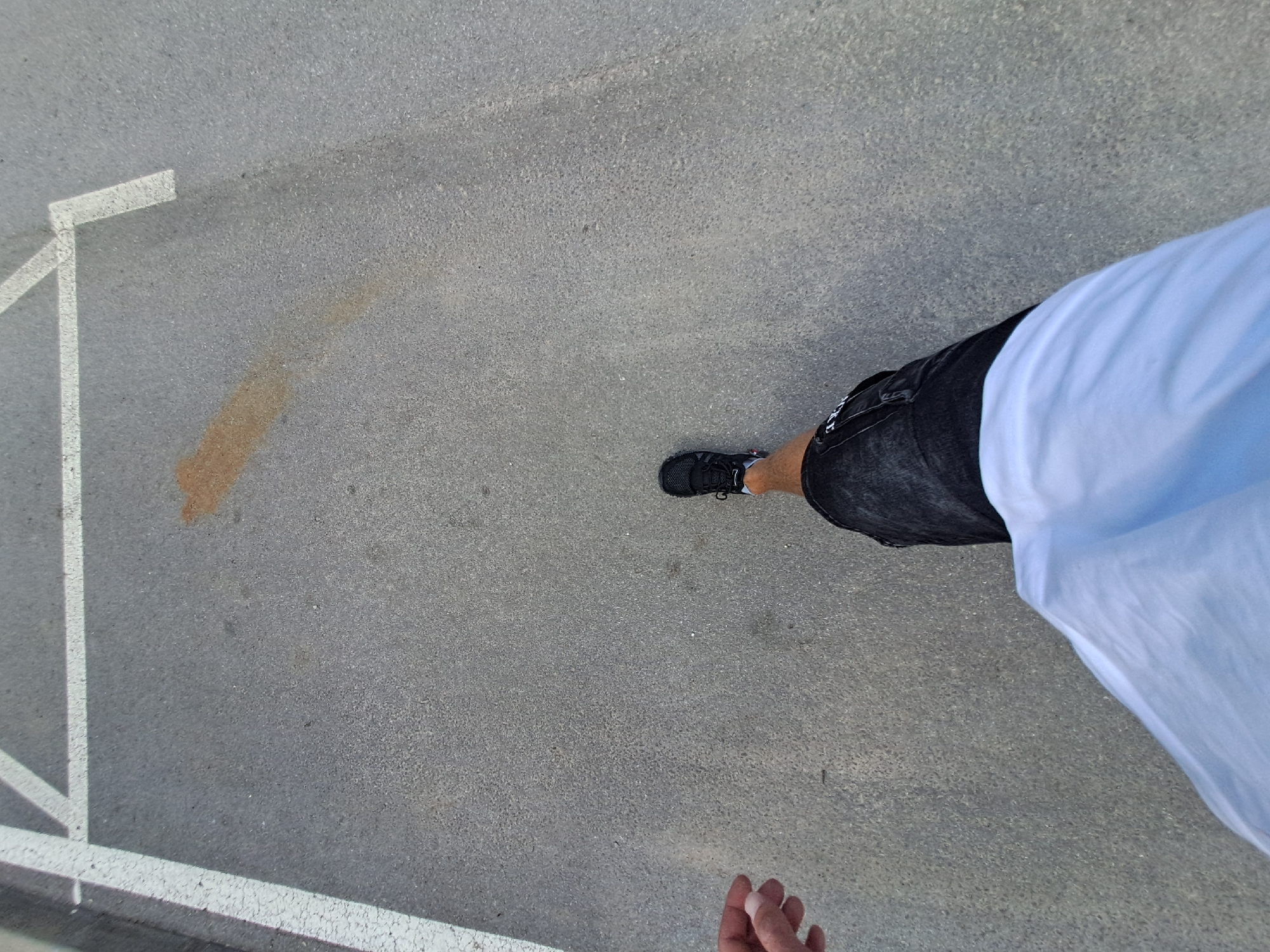 An overhead, first-person perspective shot shows a person walking across an asphalt surface. White painted lines delineate parking spaces on the asphalt. A brown stain is visible on the asphalt to the left of the walker's path. The walker wears black sneakers with white accents, dark shorts, and a light blue shirt. The walker's left hand is visible in the upper left corner of the frame.