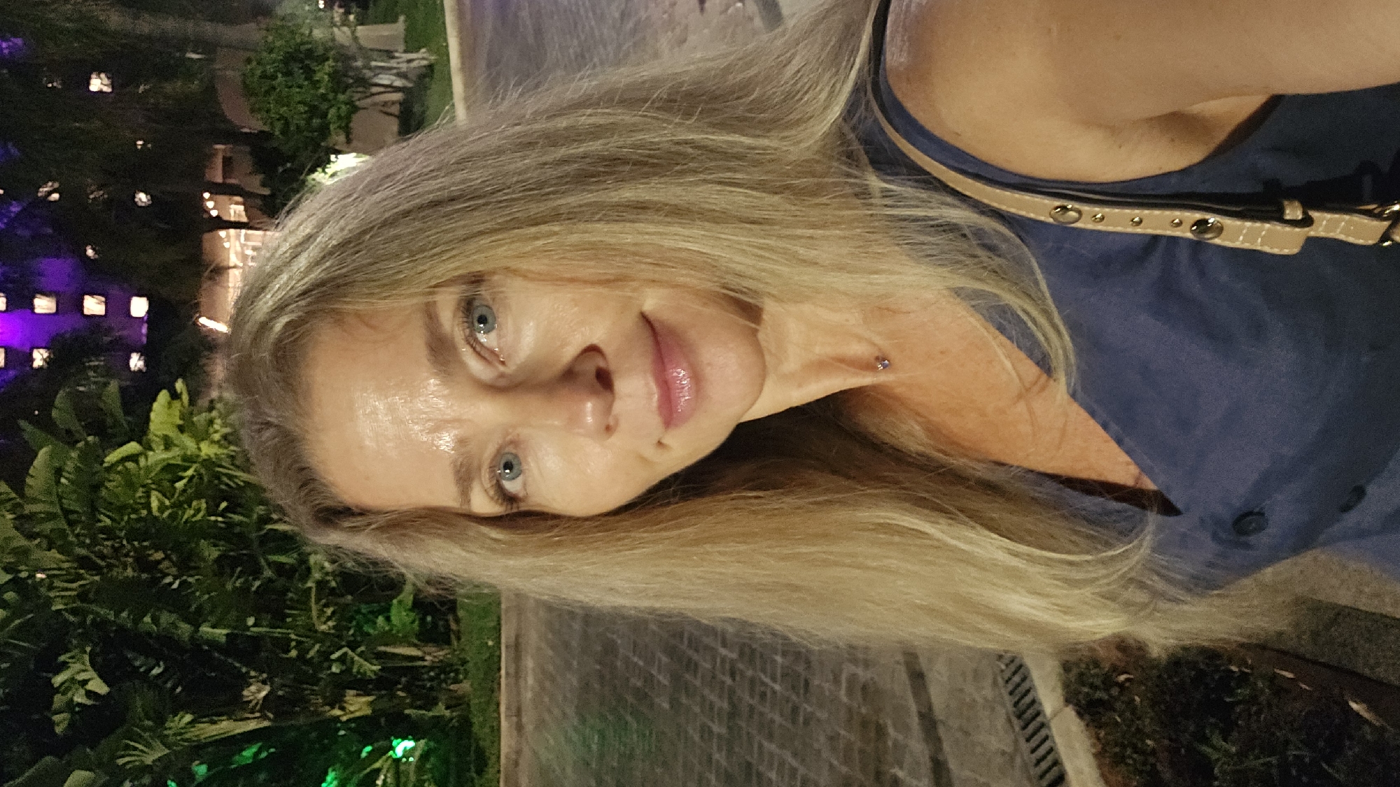 A woman with blonde hair and blue eyes is taking a selfie at night. She is wearing a blue dress and a beige purse. She is standing in front of a building with purple lights and lush green foliage. The ground is paved with cobblestones.