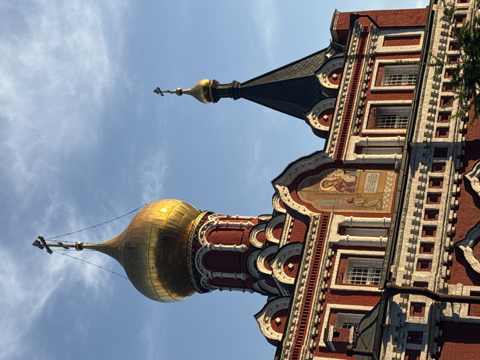 A ground-level, upward-angled shot shows the detailed upper portion of a red brick and white stone building topped with a golden onion dome and a smaller, darker spire against a blue sky. The building's architectural details include intricate stonework around windows, decorative brick patterns, and a religious mural. The golden dome reflects sunlight, and a cross sits atop both the dome and the spire. The building is located in Moscow, Russia.