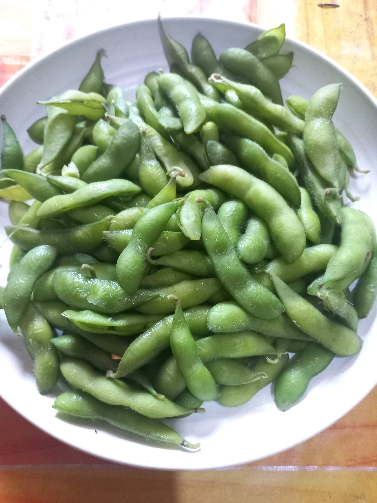 A close-up, overhead shot shows a white bowl filled with bright green edamame beans, some still in their pods and others shelled. The beans are piled high, with the light reflecting off their smooth surfaces, giving them a slightly glossy appearance. The bowl is placed on a wooden surface, which is partially visible in the foreground and background. The background of the bowl shows a subtle patterned surface. There are no people or animals present in the image, and no action or interaction is depicted. The scene appears to be a simple, still-life presentation of food. The city of Bondo, Indonesia is not depicted in this image.