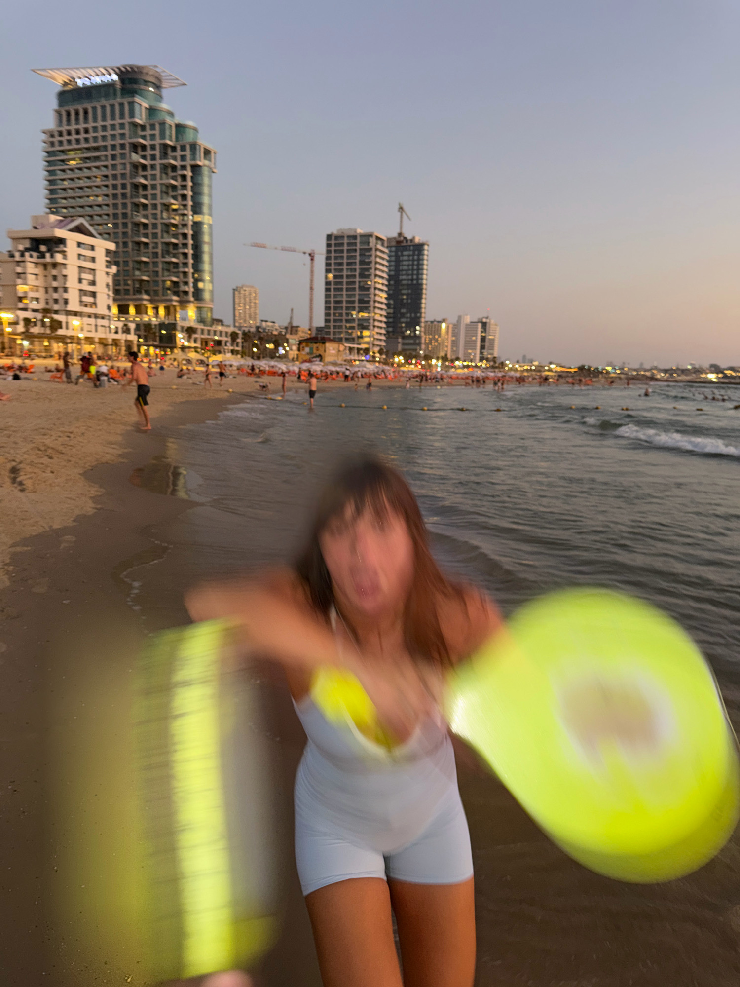 The image shows a crowded beach in Tel Aviv, Israel, at dusk. In the foreground, a young woman is blurred due to motion, holding a bright yellow frisbee. She is wearing a light blue athletic outfit. The background features the Tel Aviv coastline with several tall buildings visible, including the "Carlton Tel Aviv" hotel, and a number of other modern high-rise structures. The beach is filled with people, some are walking along the shore, others are in the water. The sea is calm with small waves. The lighting suggests the sun has recently set, casting a warm glow over the scene.