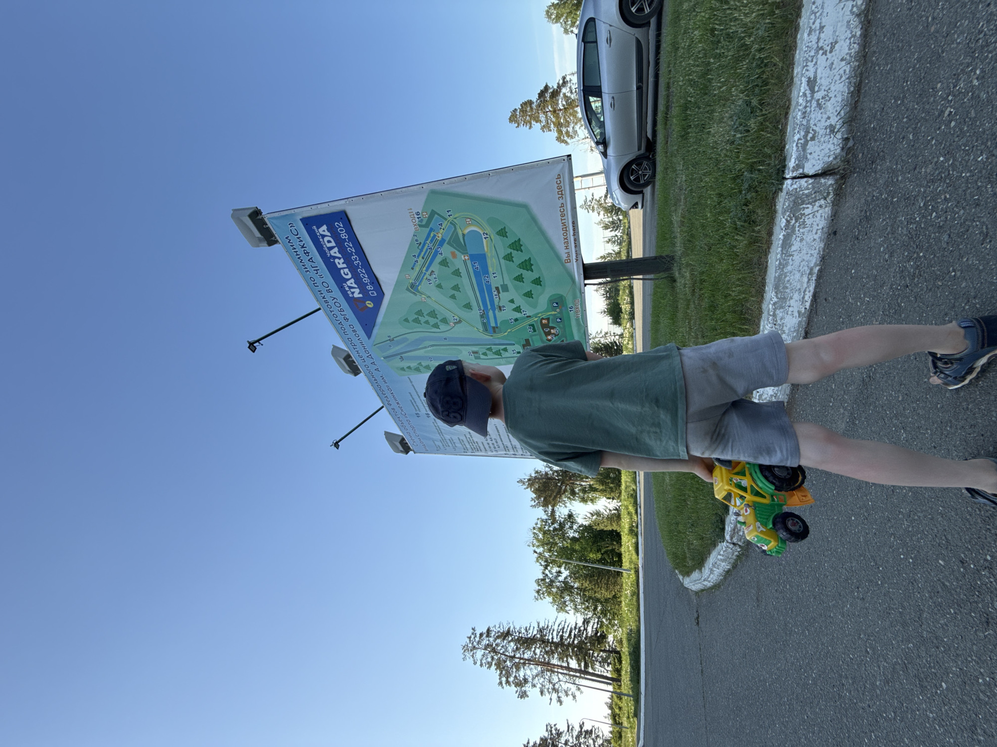A boy stands looking at a large sign in what appears to be a public park. The sign includes a map and information about the Federal Center for the Winter Sports Training of the Snowflake named after A. A. Danilov FGBOU VO "ChGAFKIS". The sign indicates a telephone number: 08-922-33-22-802. The sign also indicates that the location is in Chaikovsky. The park appears to be grassy, with trees in the background. A car is parked near the sign. The boy is wearing a green t-shirt, gray shorts, and a dark baseball cap. He is holding a green and yellow toy truck. He stands on a paved path with a concrete curb between the path and the grass.