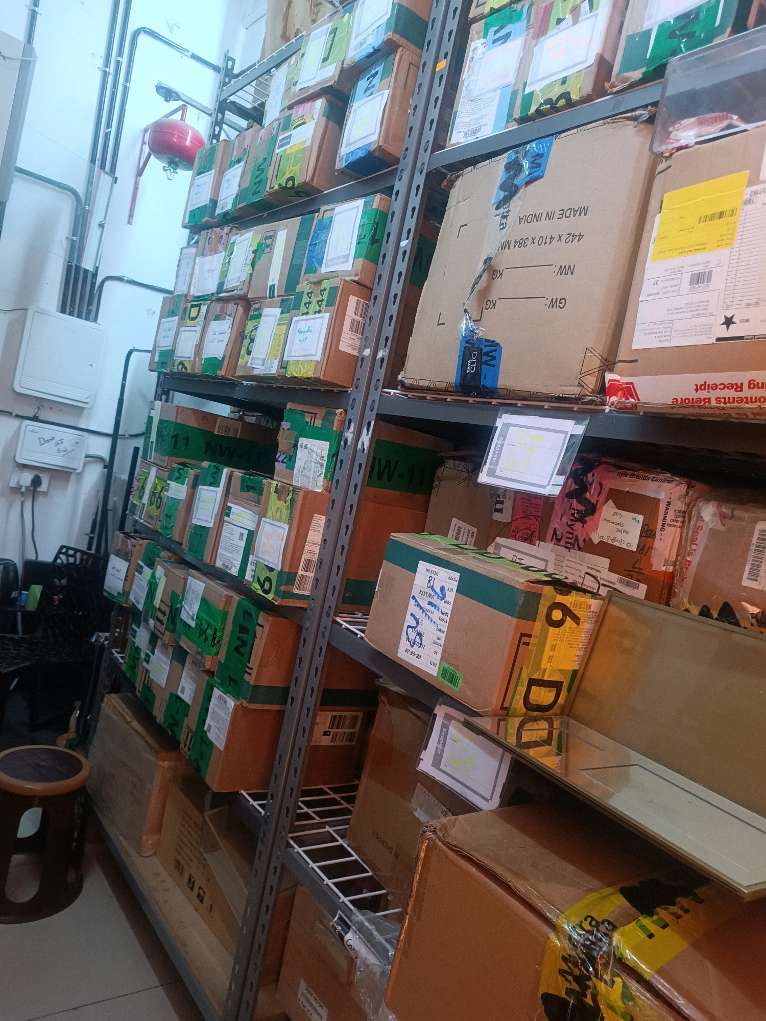The image shows a warehouse or storage facility in Hyderabad, India, filled with numerous cardboard boxes stacked on metal shelves. The shelves are organized in multiple tiers, with boxes of various sizes and designs. Some boxes are labeled with shipping information, barcodes, and text in English and what appears to be Indian languages. The lighting is artificial and casts shadows, suggesting an indoor environment. There are no people visible in the image. The overall scene depicts a well-organized but packed storage area.