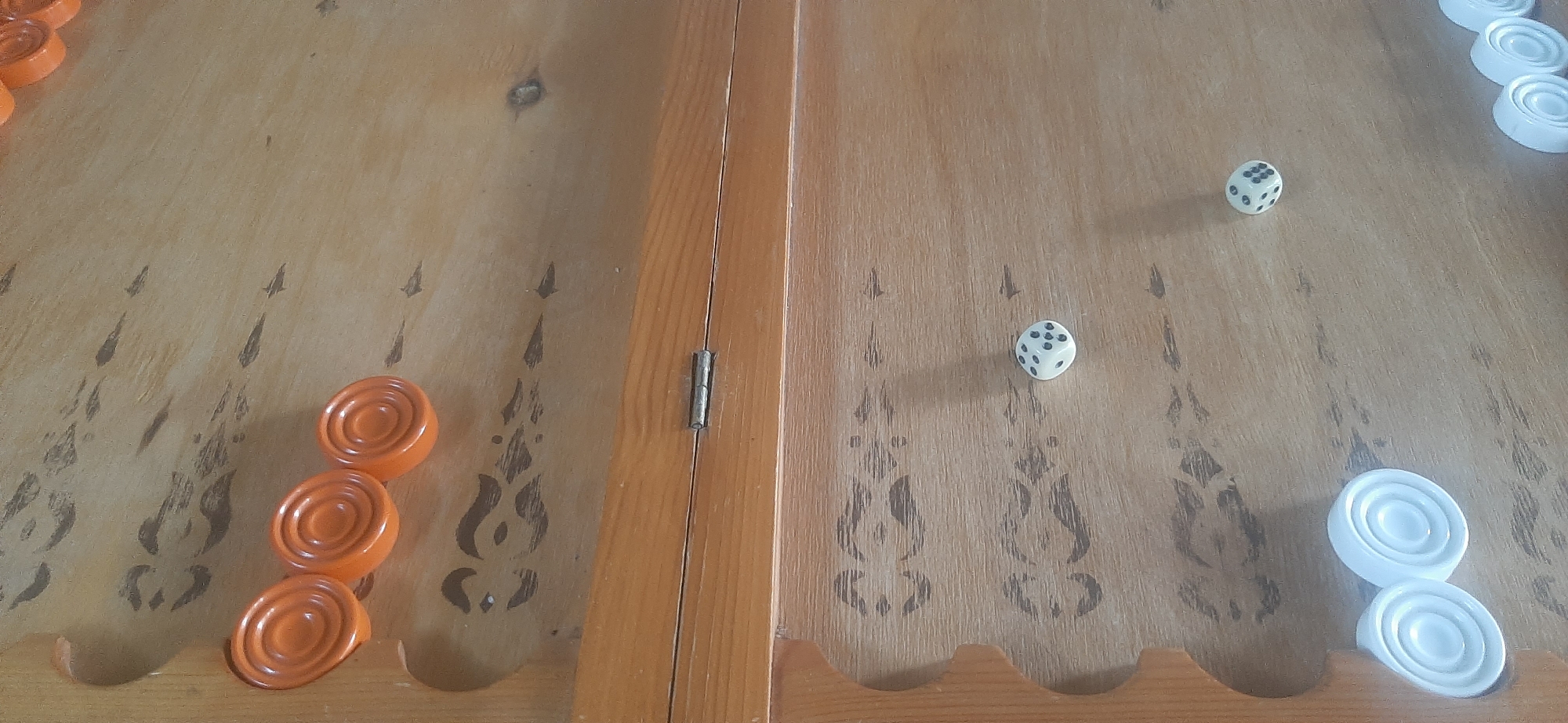 This media file depicts a close-up, overhead view of a wooden backgammon board. The board is divided by a central hinge, suggesting it can be folded. The surface of the board has a light wood grain and features decorative dark markings resembling stylized flames or icicles on both halves.
On the left side of the board, there are three orange checkers stacked on top of each other. Further up, on the very edge, are parts of two more orange checkers. On the right side of the board, there are two white checkers stacked.
Also visible on the board are two white dice. One die shows a '5' (five dots) and the other shows a '6' (six dots). The dice are positioned on the right side of the board, near the white checkers. The lighting suggests it is daytime, with a soft shadow cast by the die showing a '5'.
The scene appears to be set indoors, given the lighting and the nature of the object. There are no people visible in the media file. The image focuses solely on the game board and its pieces, implying a game is either in progress or has recently been played. The location context provided is Yessentukskaya, Russia, though there are no specific visual cues in the image to confirm this location.