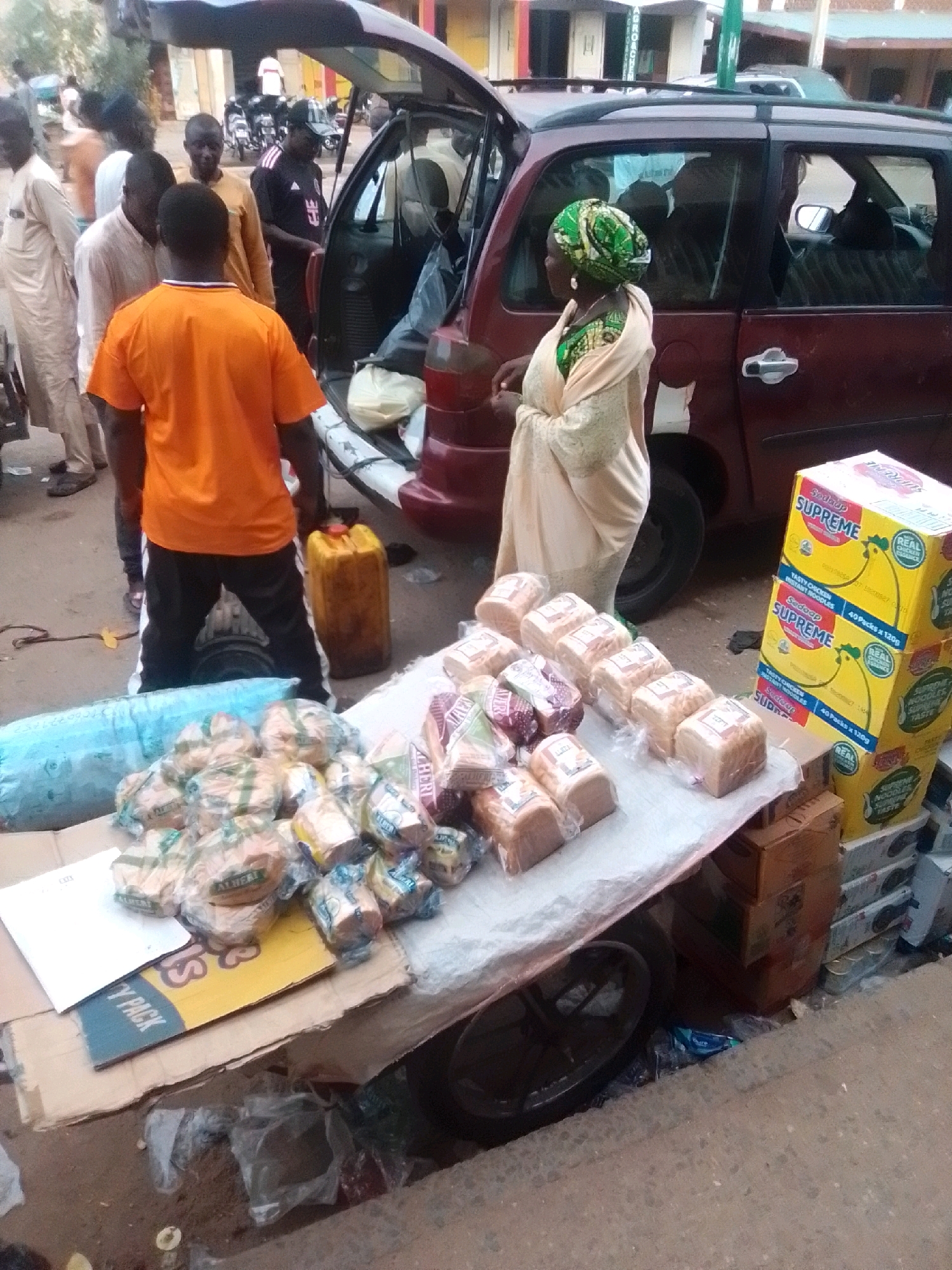 In Arawa, Nigeria, on a dusty street, a group of people are gathered around a maroon van. A woman, wearing a green headscarf and a light-colored robe, stands near the open back of the van. In front of her, a table is laden with loaves of bread packaged in plastic. To the right of the table, stacked yellow boxes with the "Supreme" logo are visible, indicating instant noodles. A man in an orange shirt stands behind the table, observing. A large yellow fuel container is placed on the ground near him. The scene suggests a bustling marketplace or roadside vending operation, with the van likely used for transportation of goods. The environment appears to be a typical urban street in Nigeria, with concrete sidewalks and buildings in the background.