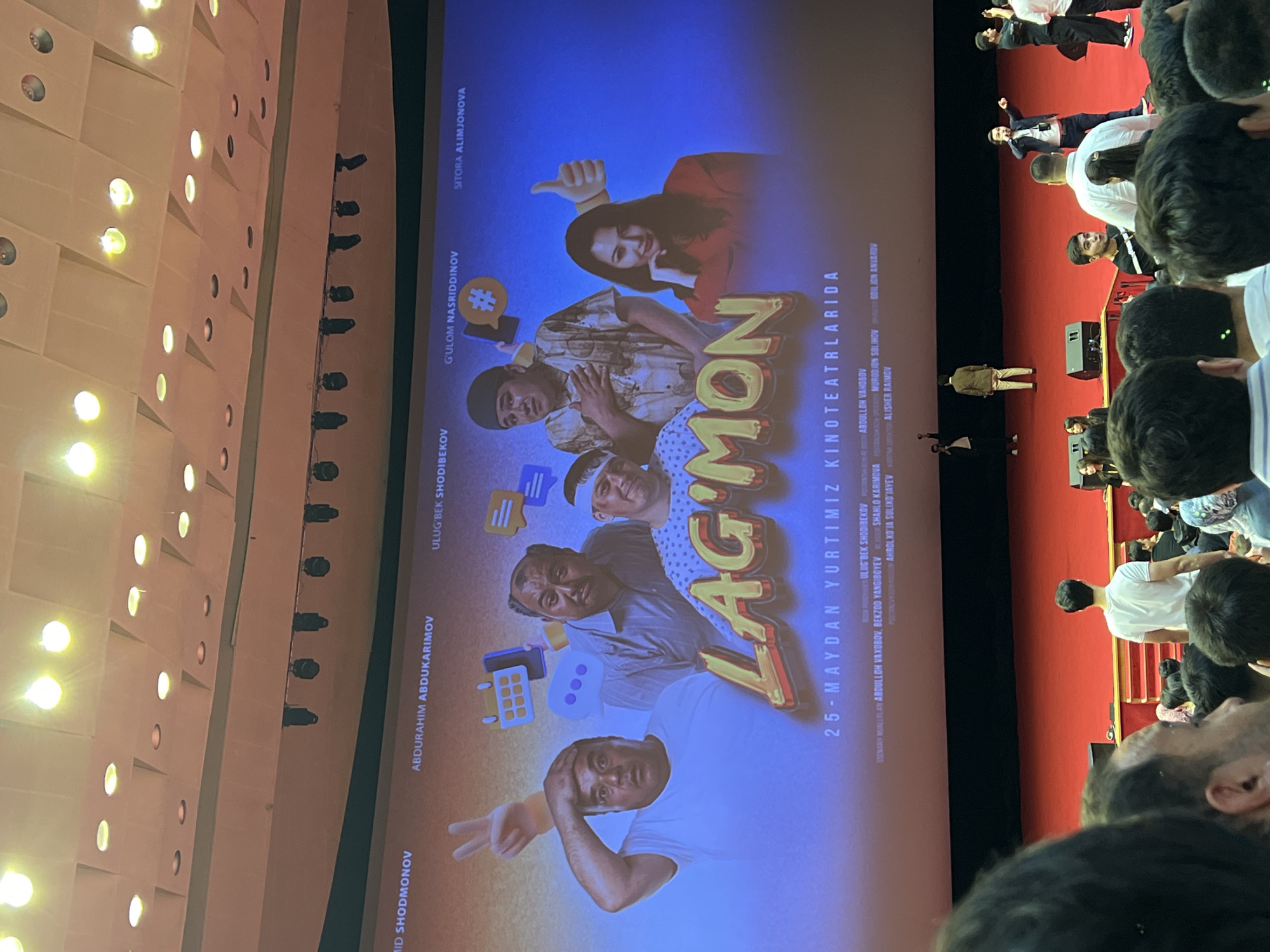The image captures a screening of the Uzbek film "Lag'mon" in a theater, likely located in Tashkent, Uzbekistan, based on the Uzbek language used in the movie poster. The audience fills the foreground, many are looking toward the screen. The poster is visible on the screen, showcasing the actors and movie title "Lag'mon". The date "25-May" suggests the film's release date. The theater's interior, including a red curtain and seating arrangement, is partly visible. A row of spotlights are seen on the wall. Some people are standing on the right in the image.