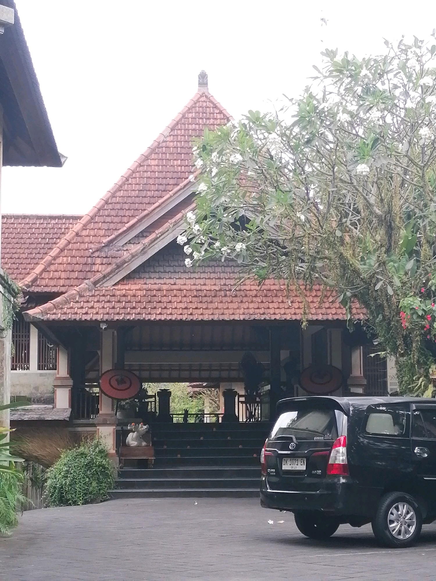 A black SUV is parked in front of a building with a red tile roof and a large covered entryway. The building appears to be a hotel or resort, with traditional Balinese architecture. There are stairs leading up to the entrance, and a statue of a white creature with orange accents sits on a wooden platform to the left of the stairs. Lush green vegetation surrounds the building and a large tree with white flowers is visible to the right. The sky is overcast.
