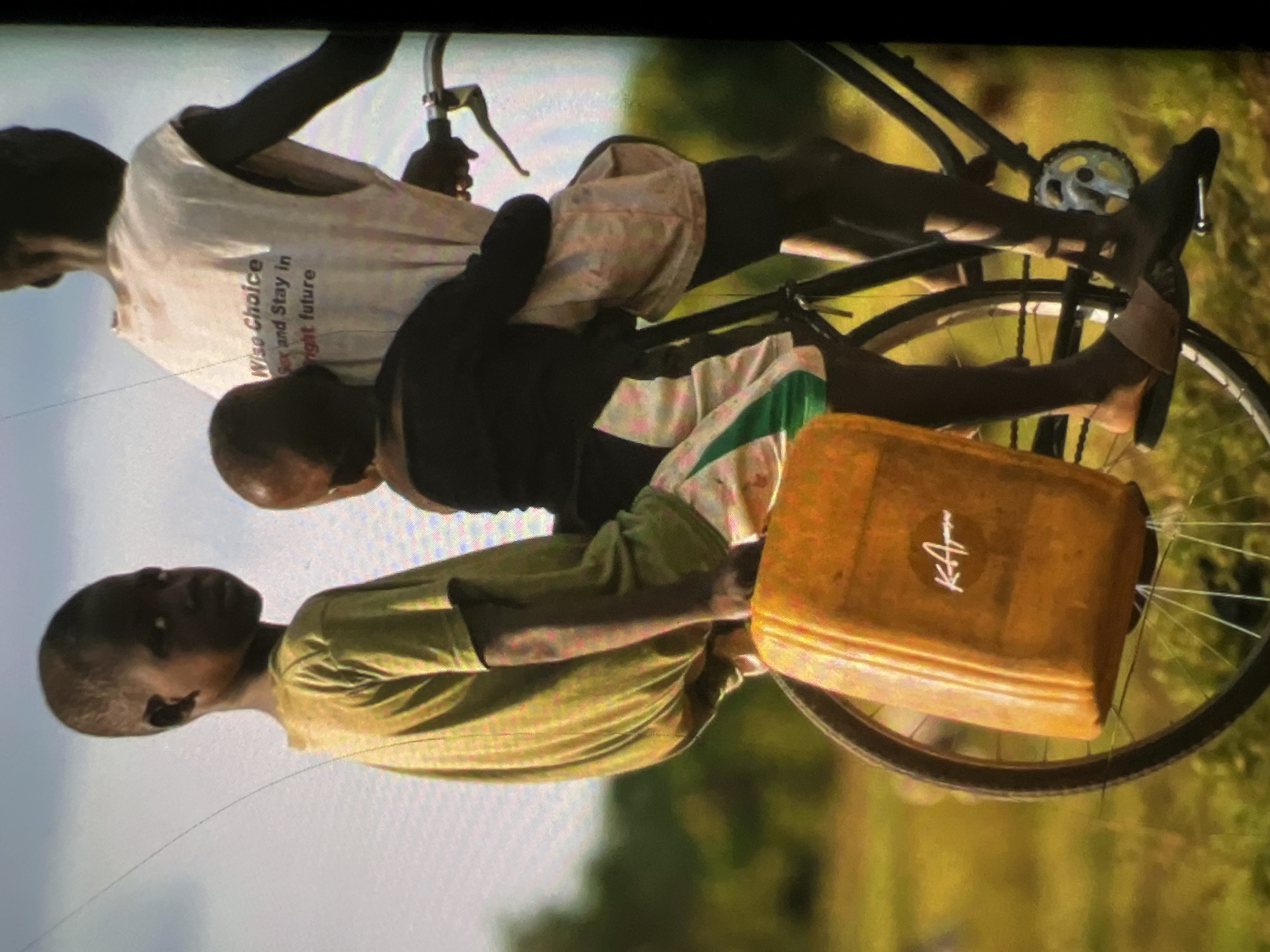 Three young boys are riding a bicycle in an outdoor, grassy environment. The boy in the front is wearing a green t-shirt and is seated on the bicycle. He is holding onto a yellow jerrycan attached to the frame of the bicycle. The second boy is sitting behind the first boy, wearing a black shirt. The third boy is riding the bicycle, wearing a white t-shirt with text that reads "Wise Choice Sex and Stay in bright future." The scene appears to be in a rural or semi-rural area, possibly a village in Lagos, Nigeria, given the context. The background is blurred, showing greenery and a light-colored sky. The overall impression is one of resourcefulness and everyday life in a developing region.