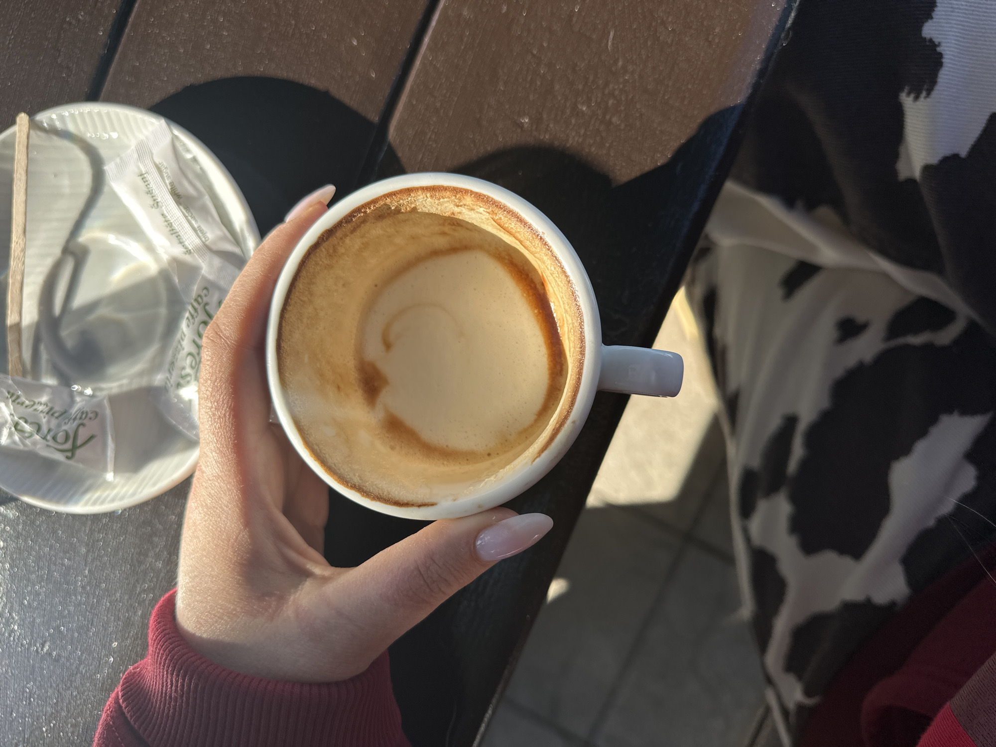 A high-angle shot shows a hand holding a half-empty white ceramic coffee cup with a light brown latte, set on a dark brown table. To the left, there's a small white saucer with a wooden stirrer and a sugar packet labeled "Fores cafe pizzerie Šušanj, Crna Gora" (Montenegro). The hand has fair skin and almond-shaped nails with a light pink polish. The person is wearing a red sweater with a black-and-white cow-print jacket. The background includes a tiled surface and the table, indicating an outdoor cafe setting in Šušanj, Montenegro. The scene is lit by bright sunlight.
