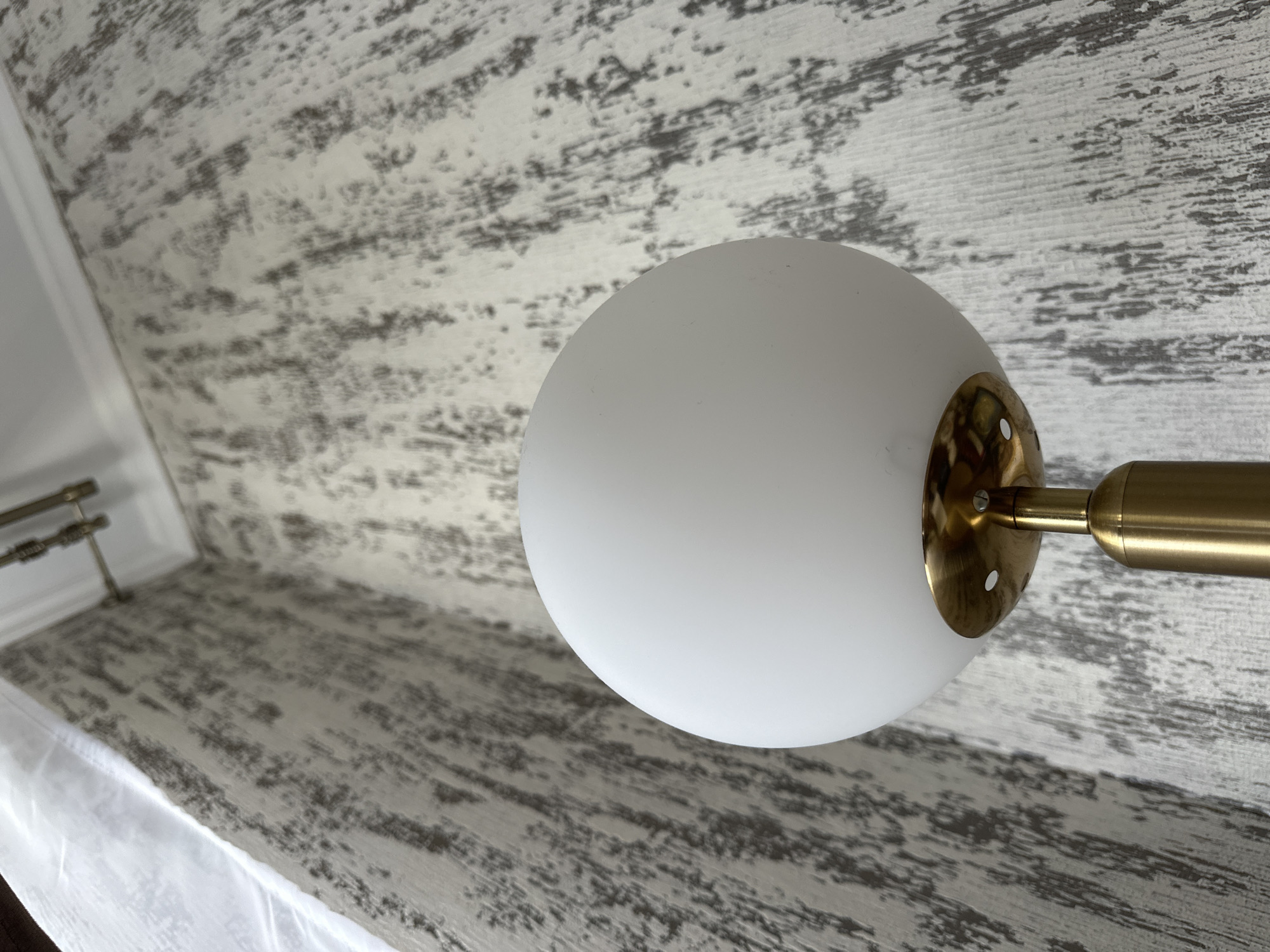 The image shows a close-up of a modern floor lamp with a white globe shade and a brass stem, set against a textured grey and white wallpaper. The wallpaper has a distressed, abstract pattern. A sheer white curtain is visible on the left side of the frame, and the corner of a ceiling with crown molding and a curtain rod is visible at the top. There are no people or animals present. The scene appears to be indoors, in a residential or commercial setting. The image was captured in Yessentuki, Russia.
