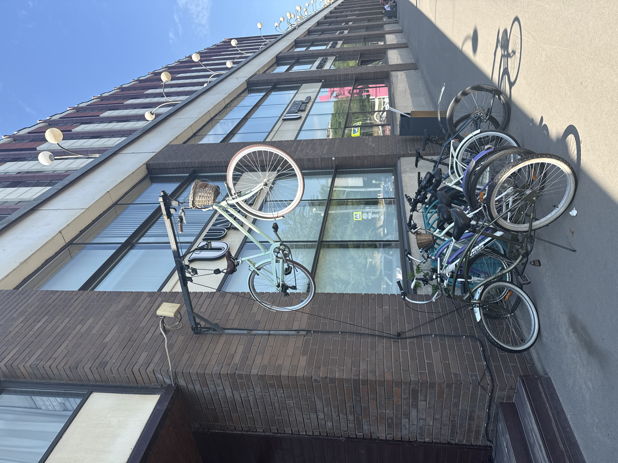 A light blue bicycle with a wicker basket is suspended by its front wheel from a metal bracket attached to the brick facade of a building. Below the suspended bicycle, a row of five bicycles are parked on a sidewalk. The building has large windows and is decorated with vertical red and white stripes on its upper section, with several globe-shaped lamps extending from its facade. The sidewalk is paved with concrete, and a shadow of a bicycle is cast on it. The sky is clear and blue. The location is the city of Zhabkino, Russia.