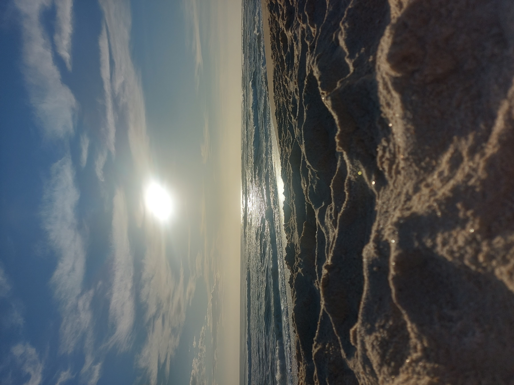 The image shows a beach scene in Figueira da Foz, Portugal. The sky, filled with wispy clouds, meets the ocean on the horizon, where the sun glares brightly. The foreground is dominated by sand, close enough to show textures and details, with scattered glints of light reflecting. The ocean has small waves.