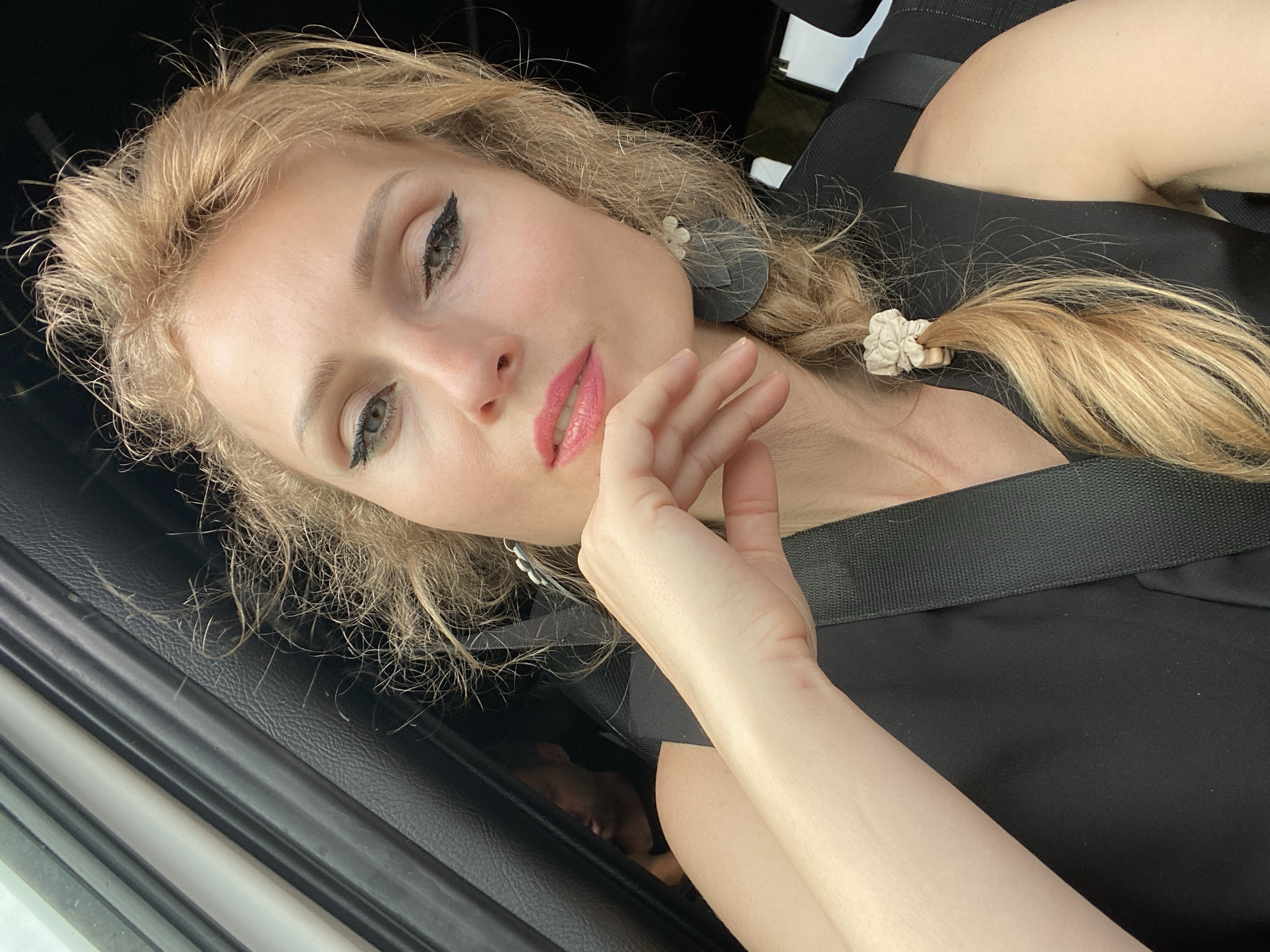 A woman with blonde hair, wearing makeup and a black top, is seen in the foreground. She has her hand on her chin and her hair is braided. A seatbelt is visible across her chest. In the background, another person is partially visible in the back seat of a vehicle, which appears to be in motion. The scene suggests the woman is a passenger in a car, possibly during daytime. The location is specified as Valozhynski selski Savet, Belarus.