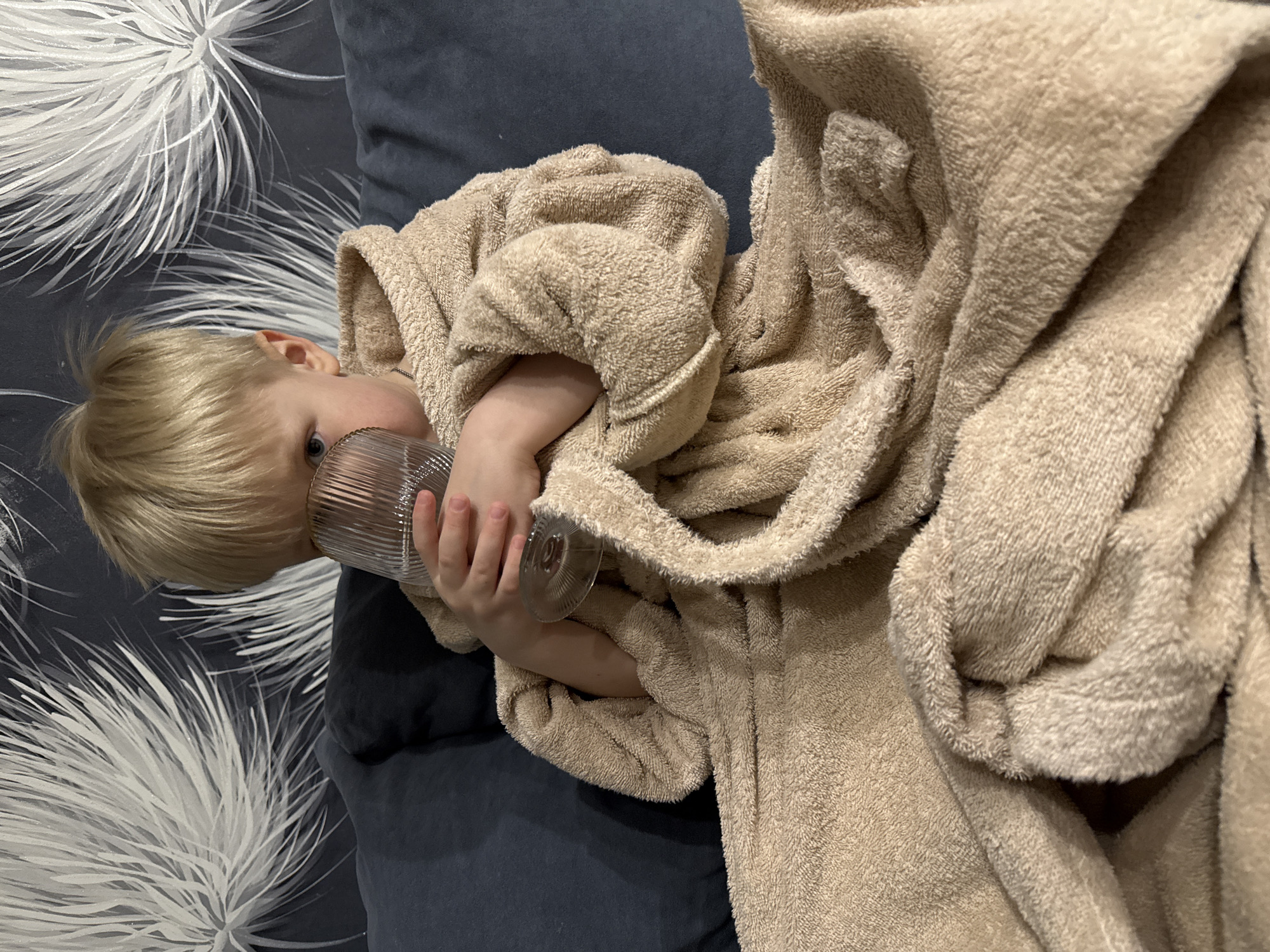 A young child with blond hair and blue eyes is depicted lying down, viewed from a slightly overhead perspective. The child is wearing a soft, beige terry cloth bathrobe, which is draped around their body and arms. With both hands, they are holding a clear glass that has a distinct ribbed or fluted texture, bringing it up towards their mouth or face.
The child appears calm and relaxed. The setting is indoors, with the child resting on a dark blue or gray surface, likely a sofa or bed. Behind them, a patterned surface, possibly a cushion or wallpaper, features white, spiky, or feathery designs against the dark background, resembling dandelions or fireworks. No text is visible in the image. This media file originates from Chaikovsky, Russia.