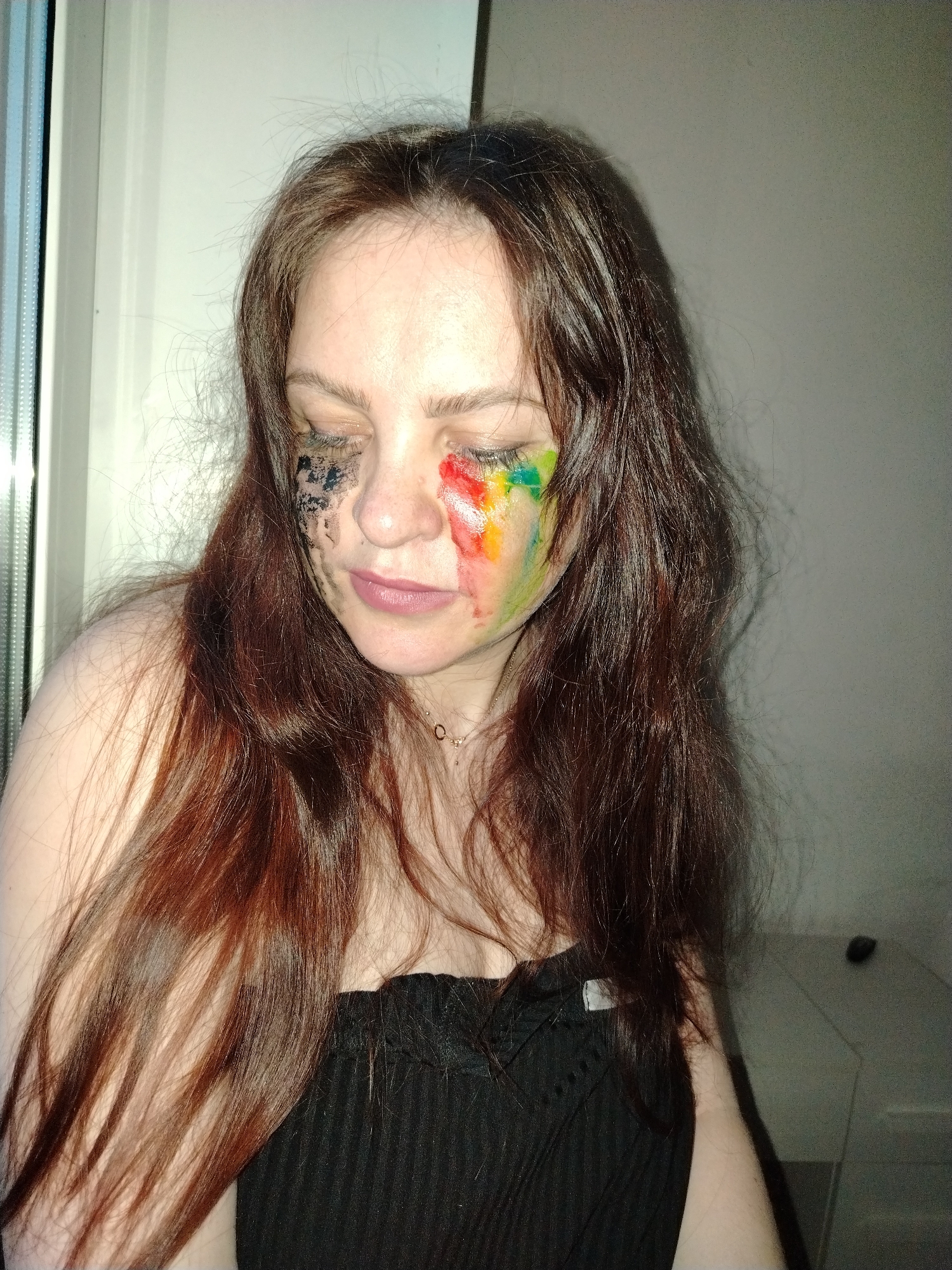 A person with light skin and long, auburn hair is depicted indoors. The person is wearing a black, strapless top. The person's face has makeup or paint on either side of their face in a manner suggesting tears. The makeup on the left side of the face is black, while the makeup on the right side consists of a rainbow color scheme. The person has their eyes closed and is looking down. A window can be seen on the left side of the image, and a wall can be seen on the right side of the image. There is no indication of this image being in the city of Noviy, Russia.
