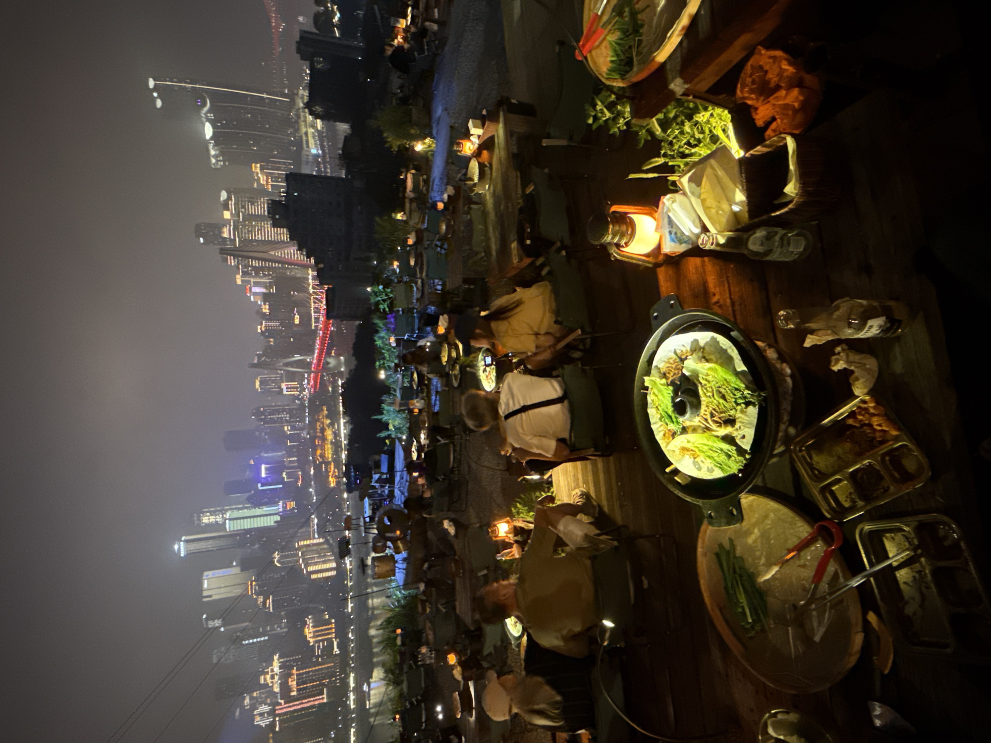 The image captures an outdoor dining scene at night, with a view overlooking the illuminated cityscape of Nan'an District, China. The perspective suggests the photo was taken from a slightly elevated position, focusing on a wooden deck area.
Several tables are occupied by diners. Each table is set with a hot pot dish, surrounded by plates and bowls containing food. Lanterns provide warm lighting on some tables. The composition places emphasis on the dining experience against the backdrop of the city lights.
The city skyline is prominently displayed with numerous skyscrapers illuminated, highlighting the urban density and architectural complexity of Nan'an District at night. The sky is dark, adding contrast and emphasizing the vibrant glow of the city below. Electrical wires are visible.